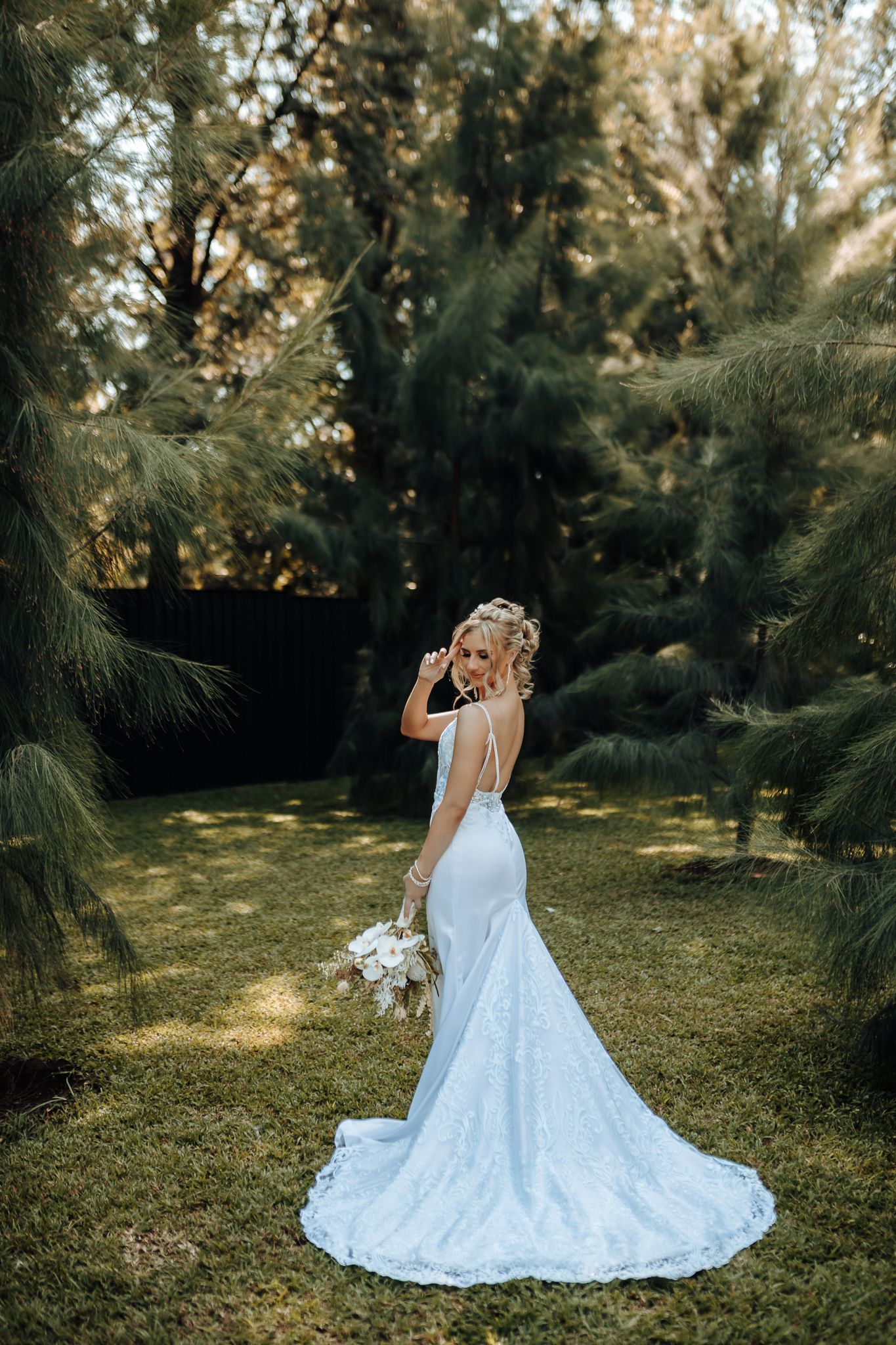 Bride in fitted lace gown with long train poses in garden with dappled sunlight.