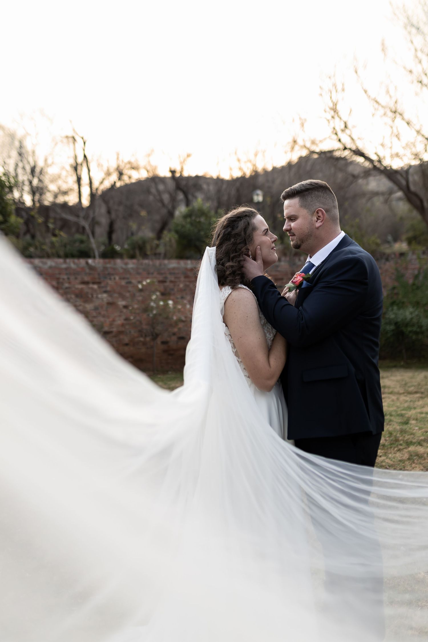 A romantic wedding couple embraces outdoors as the bride's long white veil flows dramatically in the evening light.