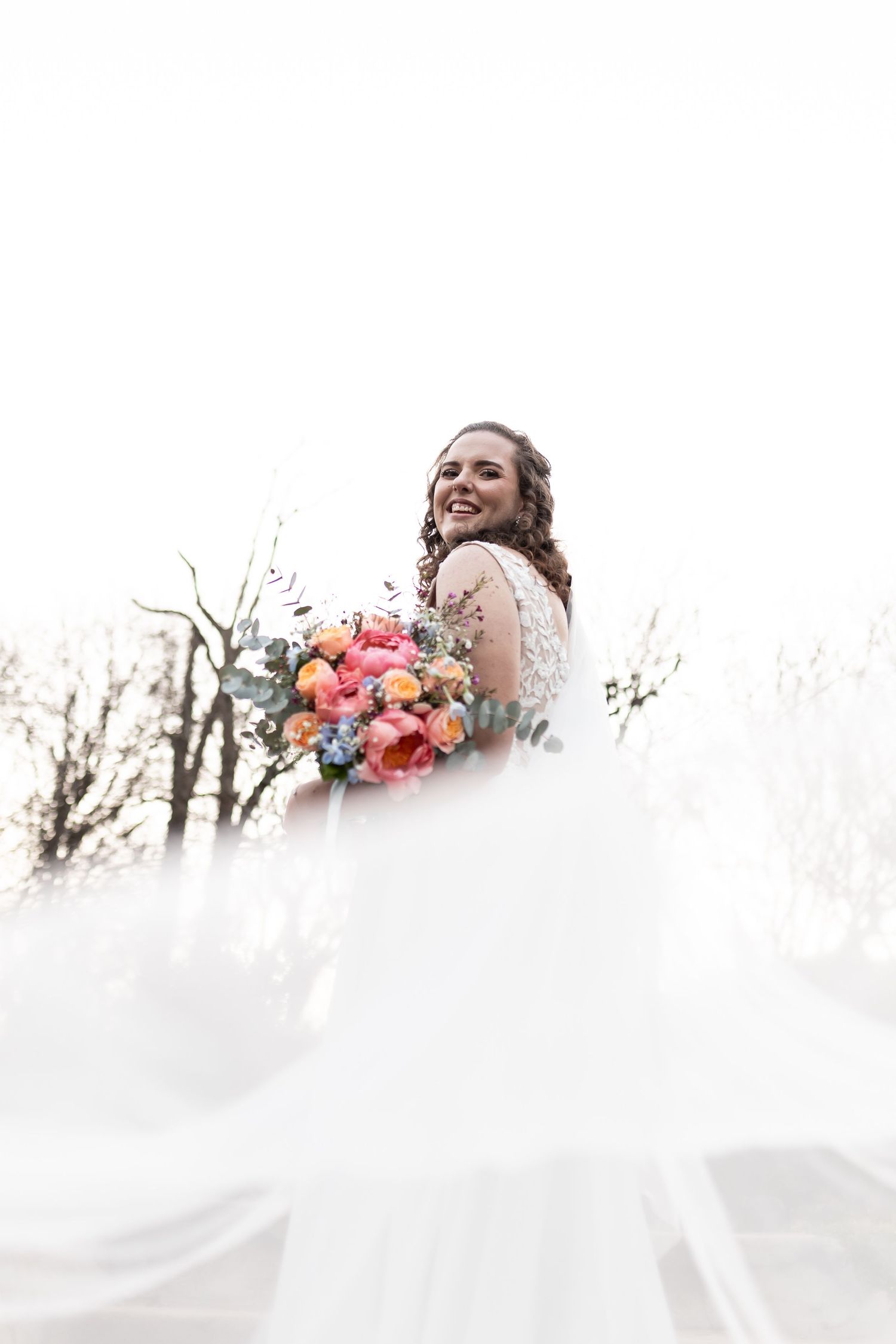 A joyful bride in a flowing white gown holds a vibrant pink and coral bouquet while her dress billows in the breeze.