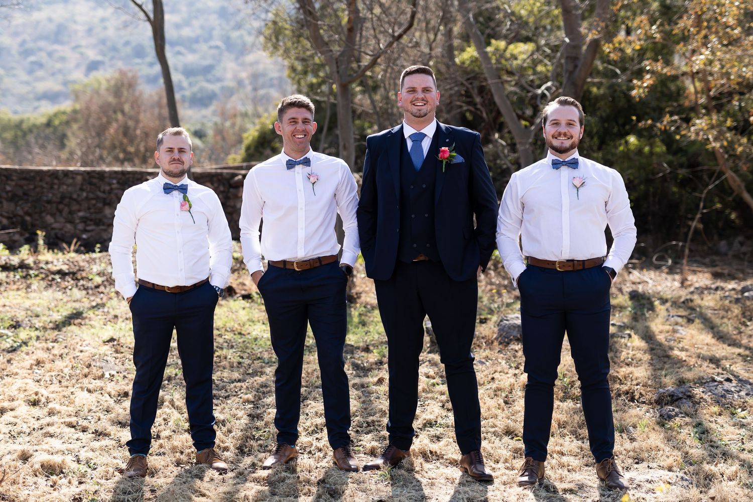 A groom and groomsmen stand together outdoors in formal attire with bow ties and suspenders on a sunny day.