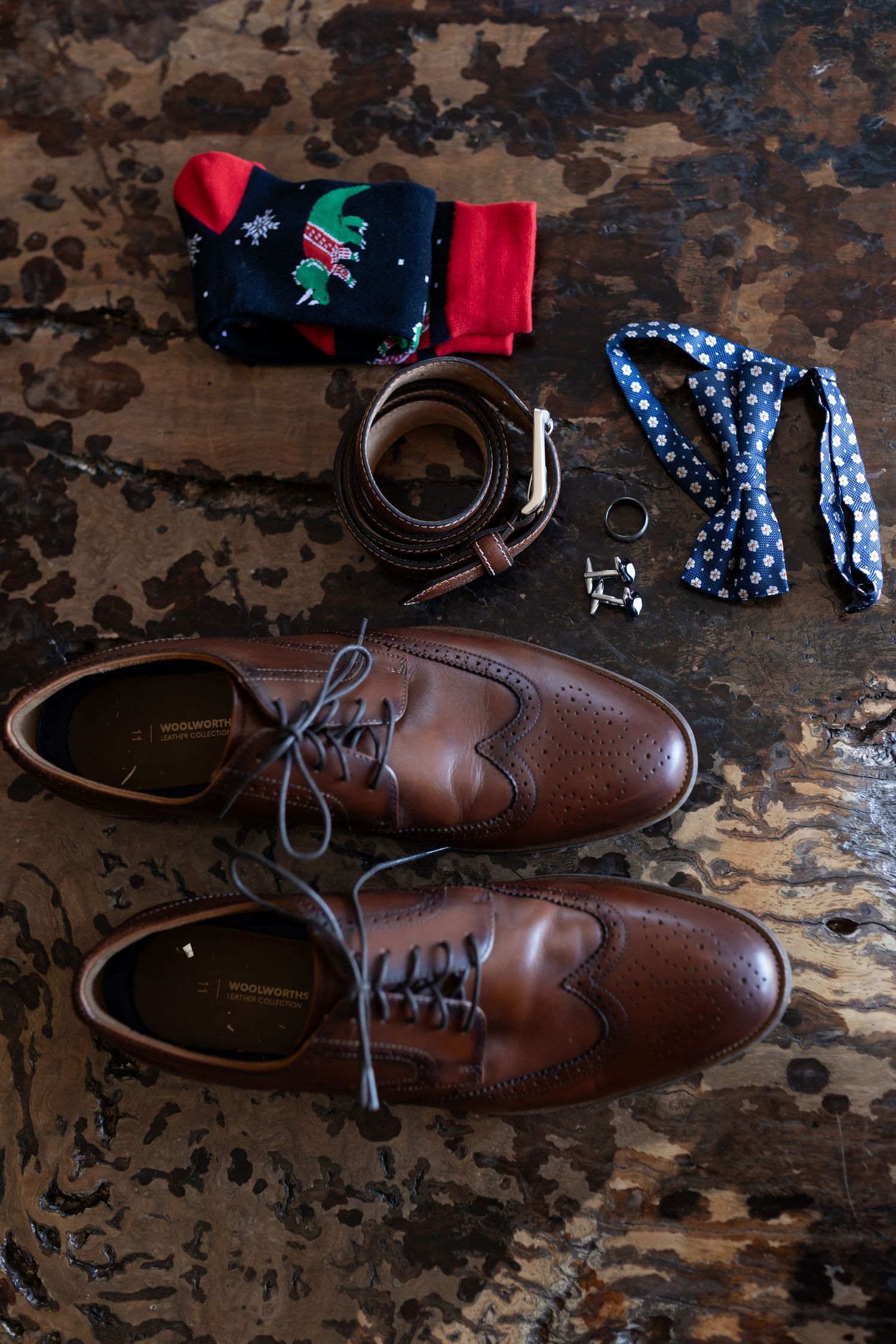 Brown leather dress shoes with Christmas socks, bow tie and accessories arranged on rustic wooden surface.