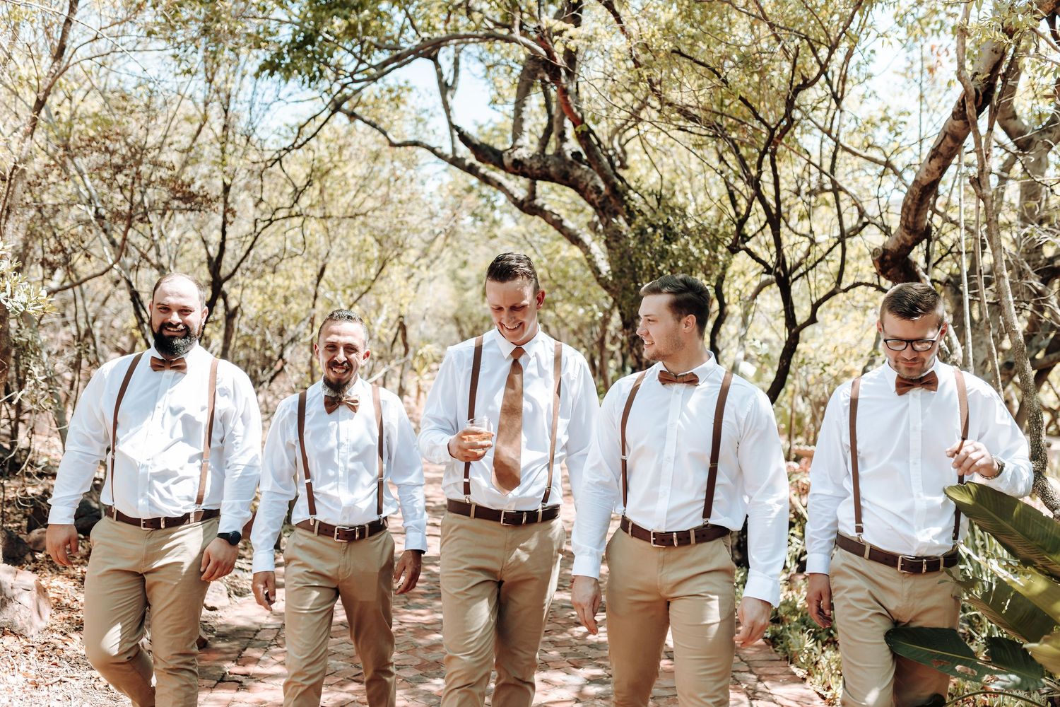 Group of groomsmen in coordinated attire walking together on wooded path at wedding.