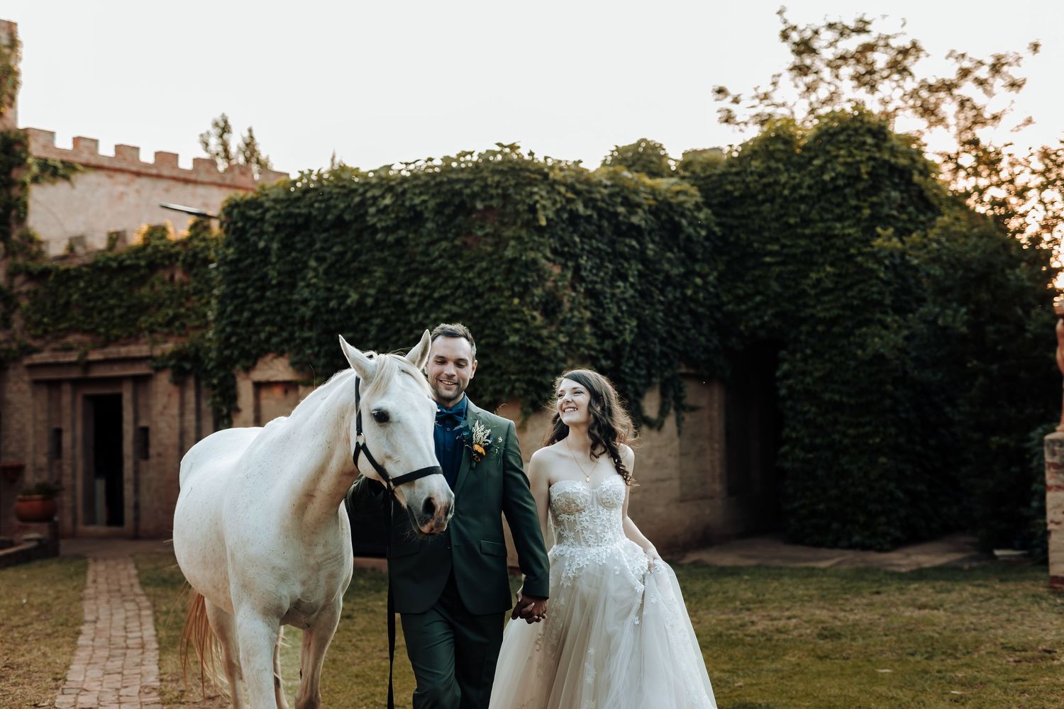 Wedding day portrait with couple and white horse in front of a historic castle building.