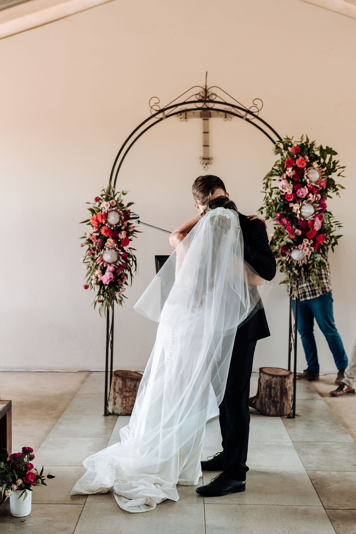 Couple stands beneath an ornate metal arch decorated with pink and red flowers during wedding ceremony.