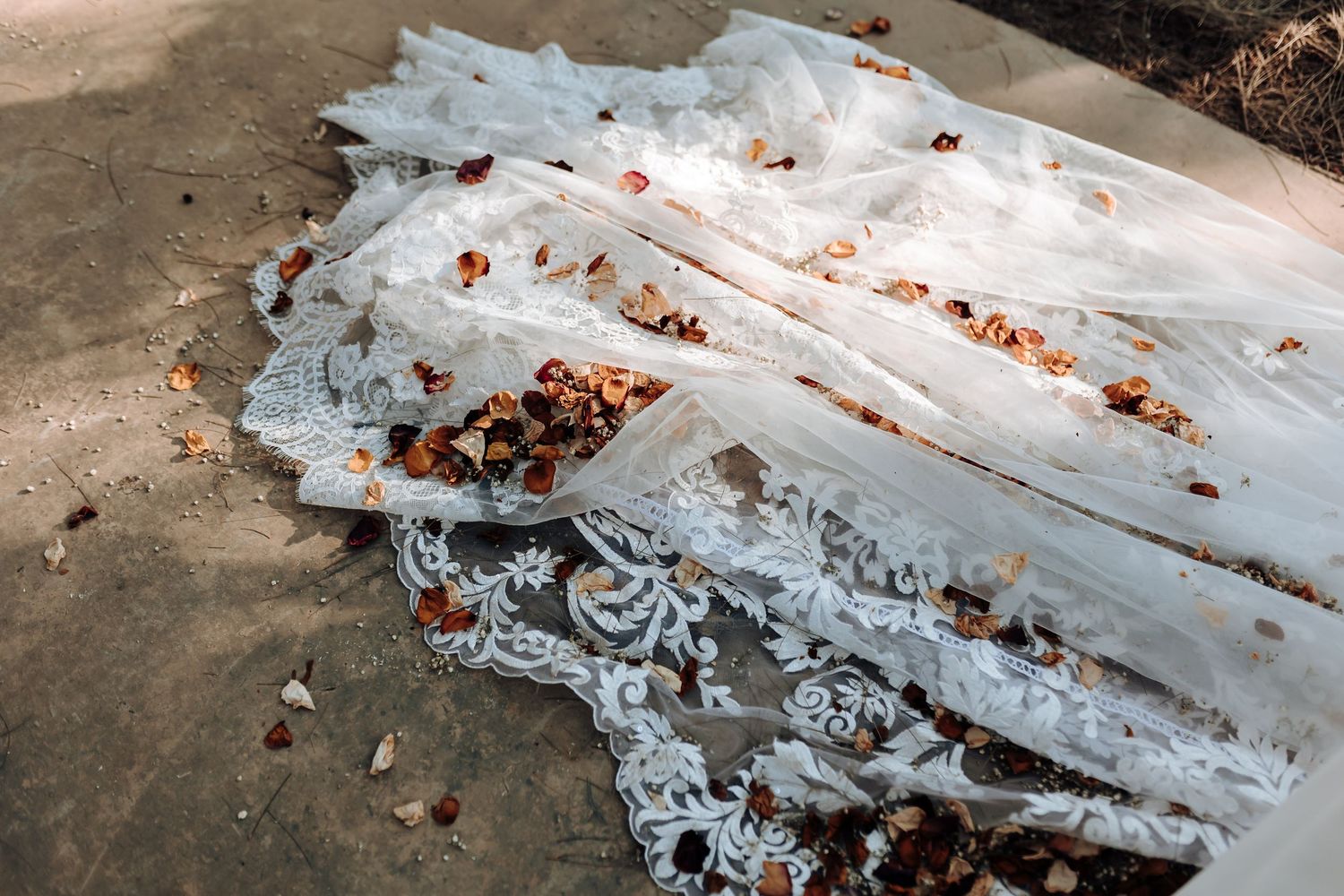 A delicate white lace wedding dress train laid out on concrete with scattered rose petals.