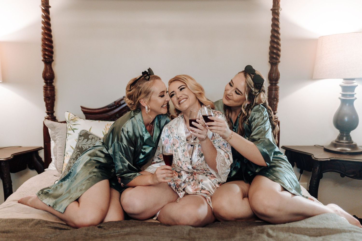 Three bridesmaids in silk robes share a joyful moment on an elegant four-poster bed during wedding preparations.