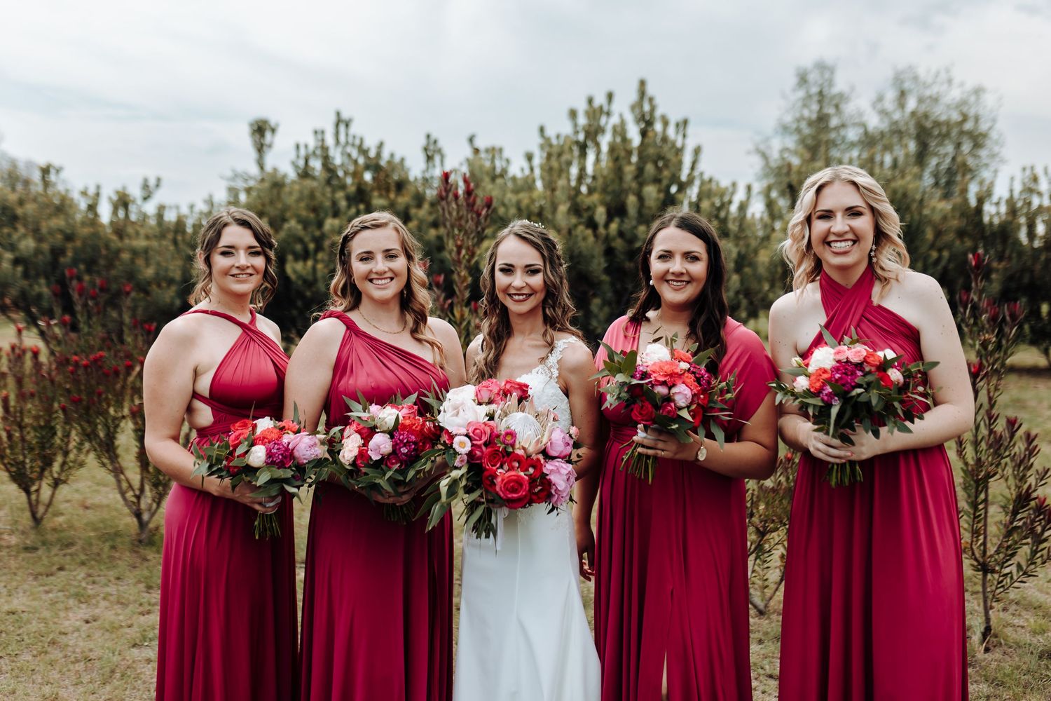 Wedding party in burgundy dresses holding matching floral bouquets outdoors.