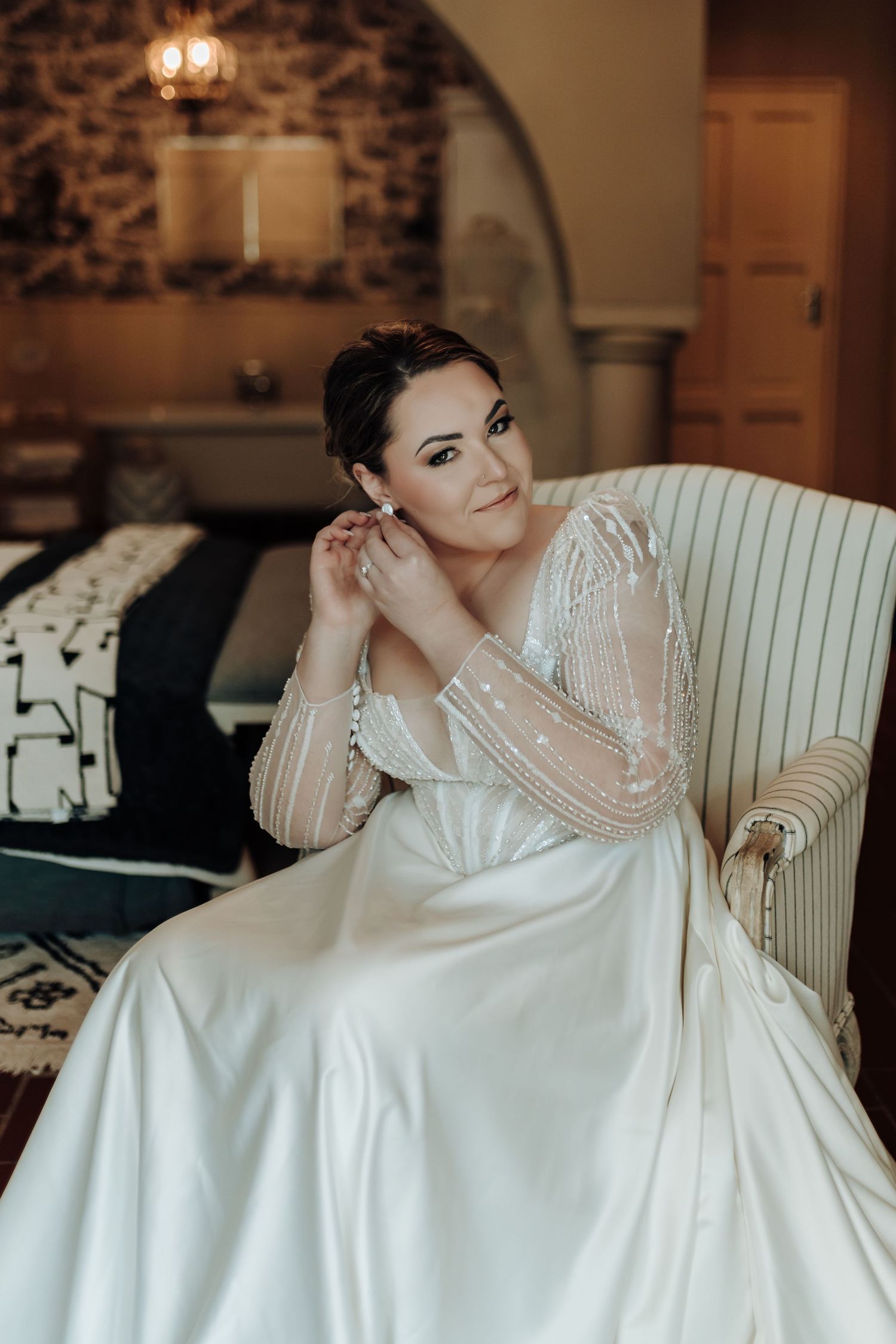 Classic bridal portrait in a white chair showing off intricate beadwork and flowing satin skirt.