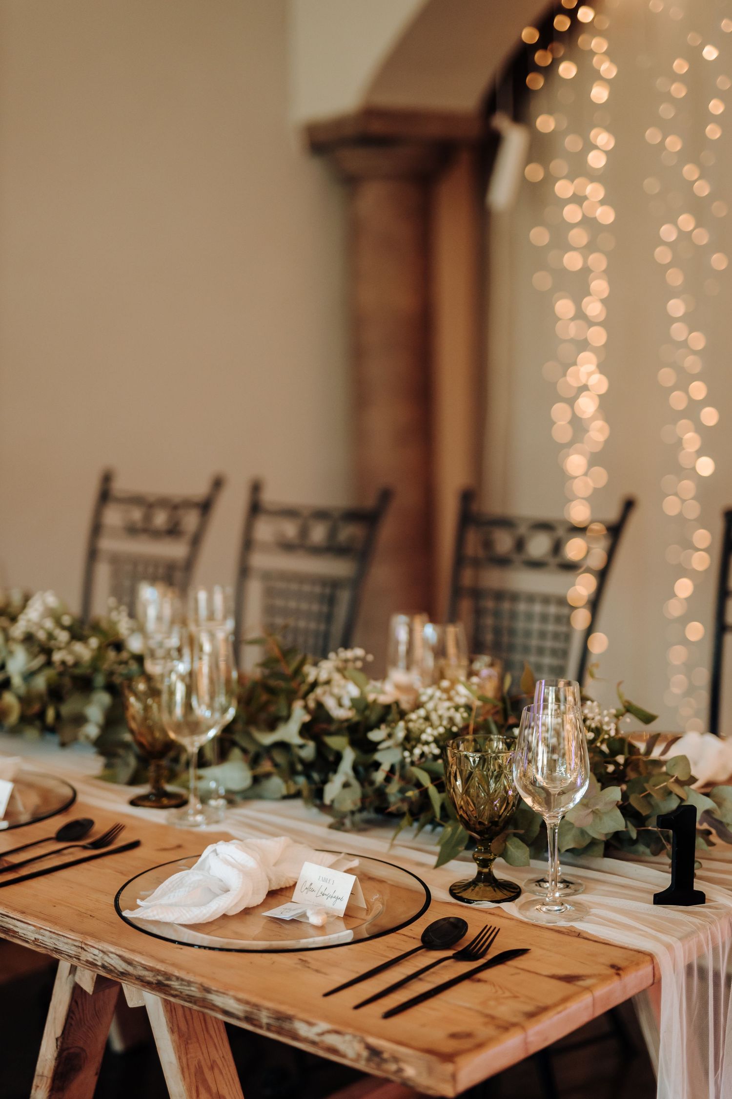 Rustic wooden table set for wedding reception with greenery garland and twinkling fairy lights.