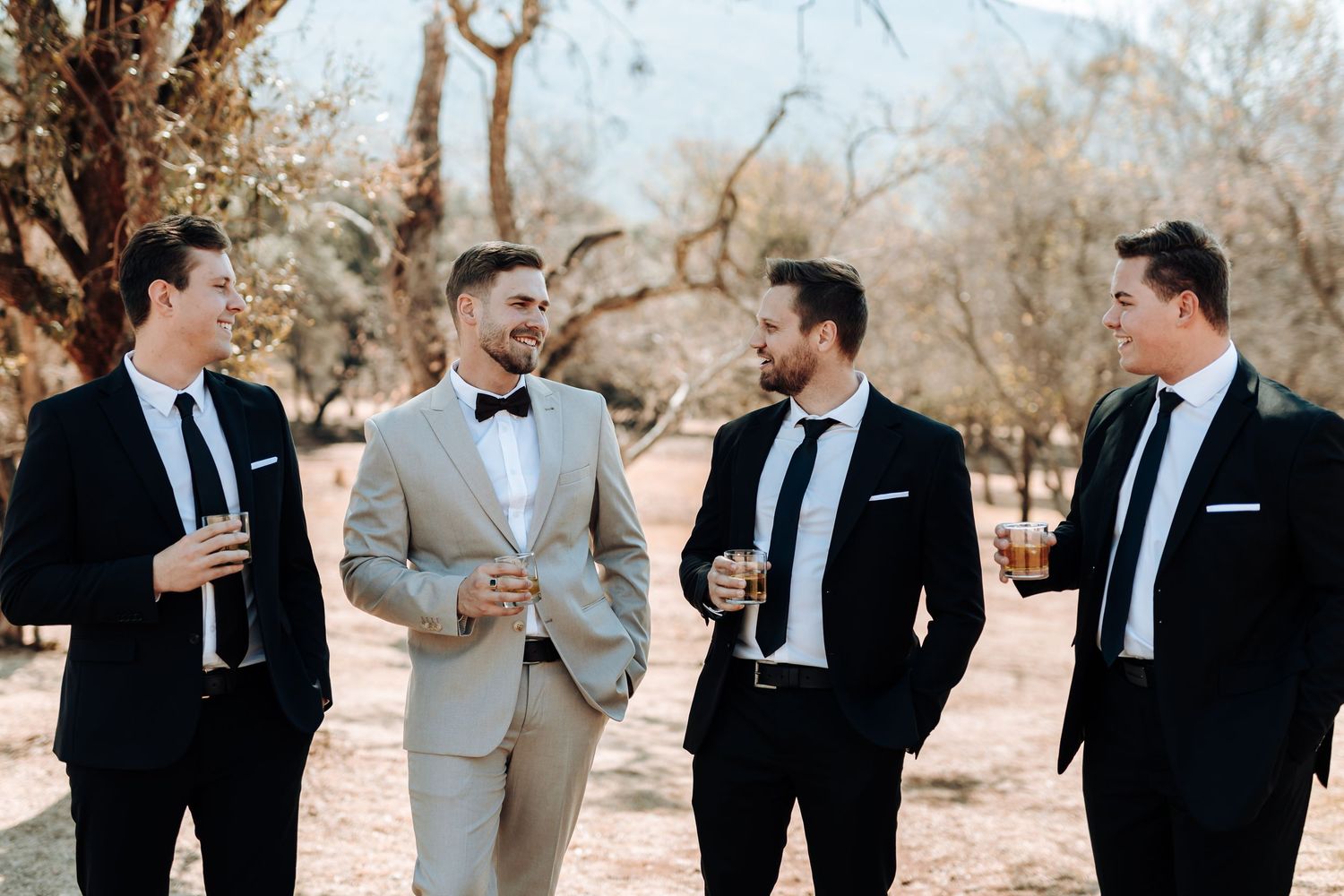 Wedding party poses outdoors among trees with groom in beige suit surrounded by groomsmen in black.
