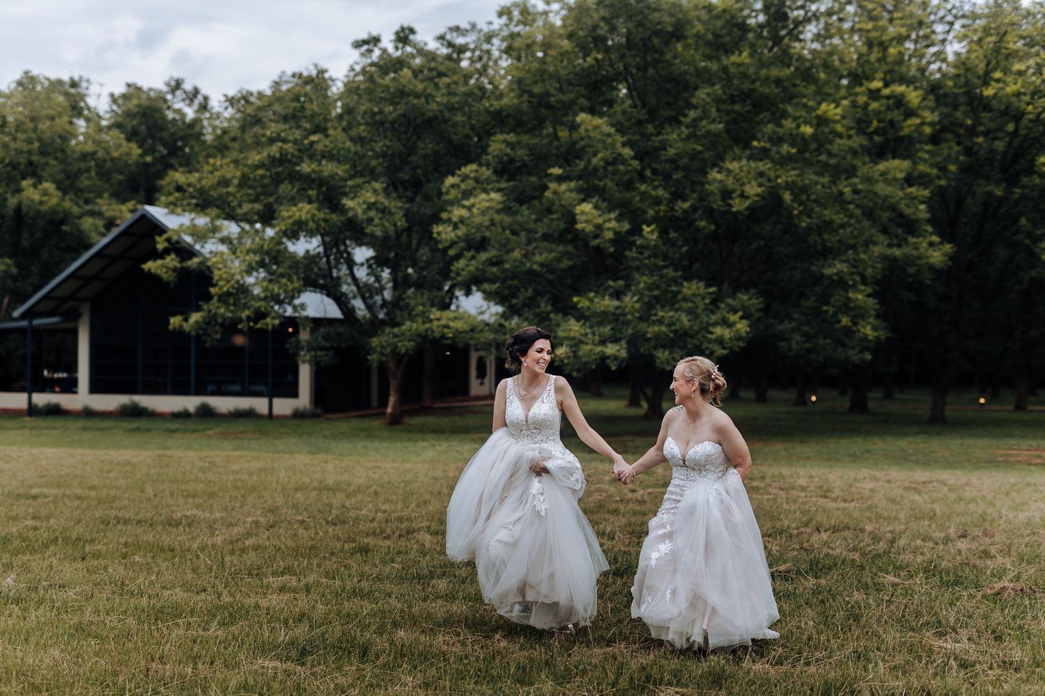 Two people in white wedding dresses running together across a grassy field on an overcast day.