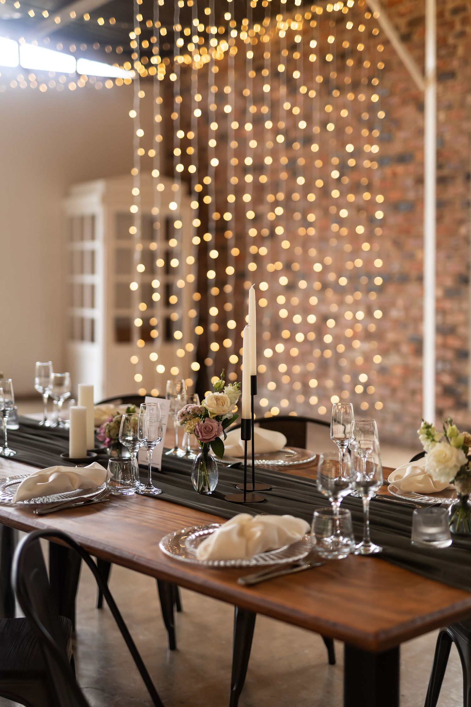 Rustic wooden table set for dining with elegant place settings and twinkling fairy light backdrop.