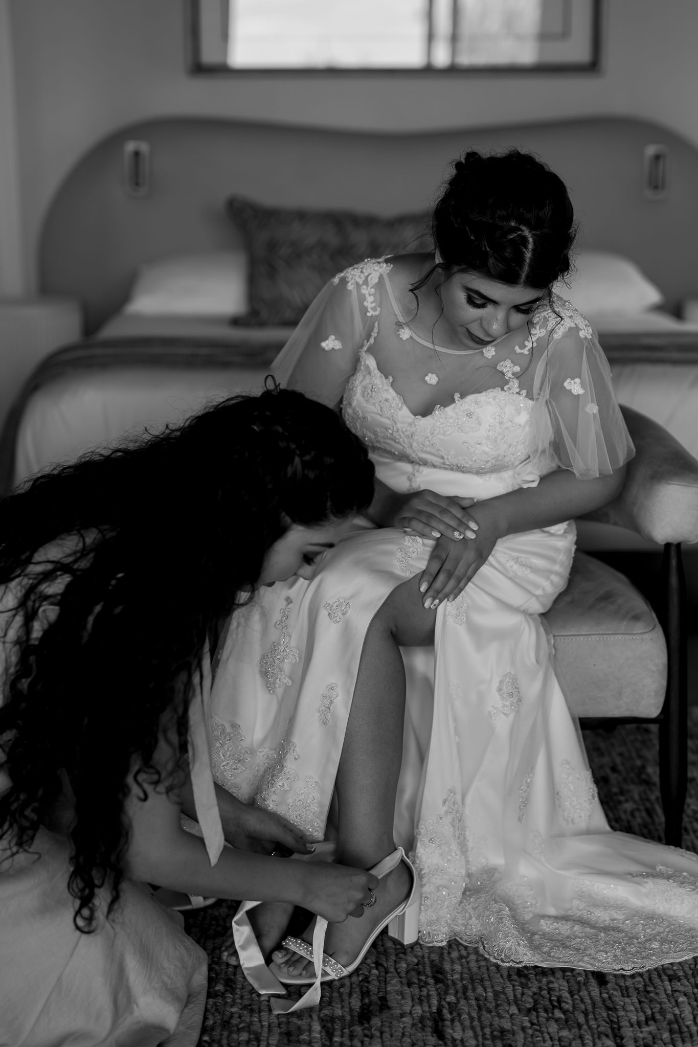 Black and white photo of wedding preparation with white lace dress and shoe fitting.