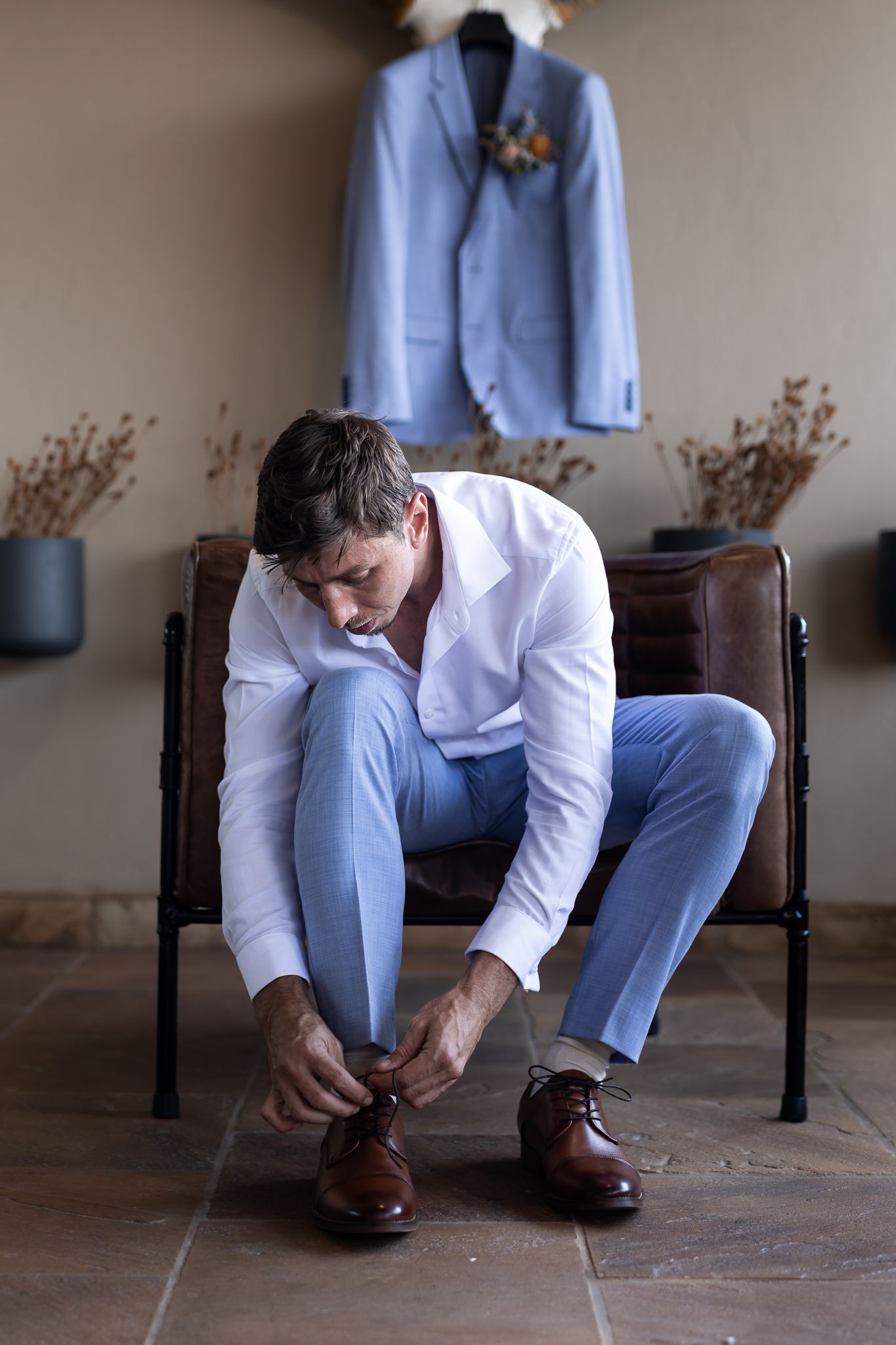 Person in white shirt and blue pants tying shoes on leather chair with suit displayed above.