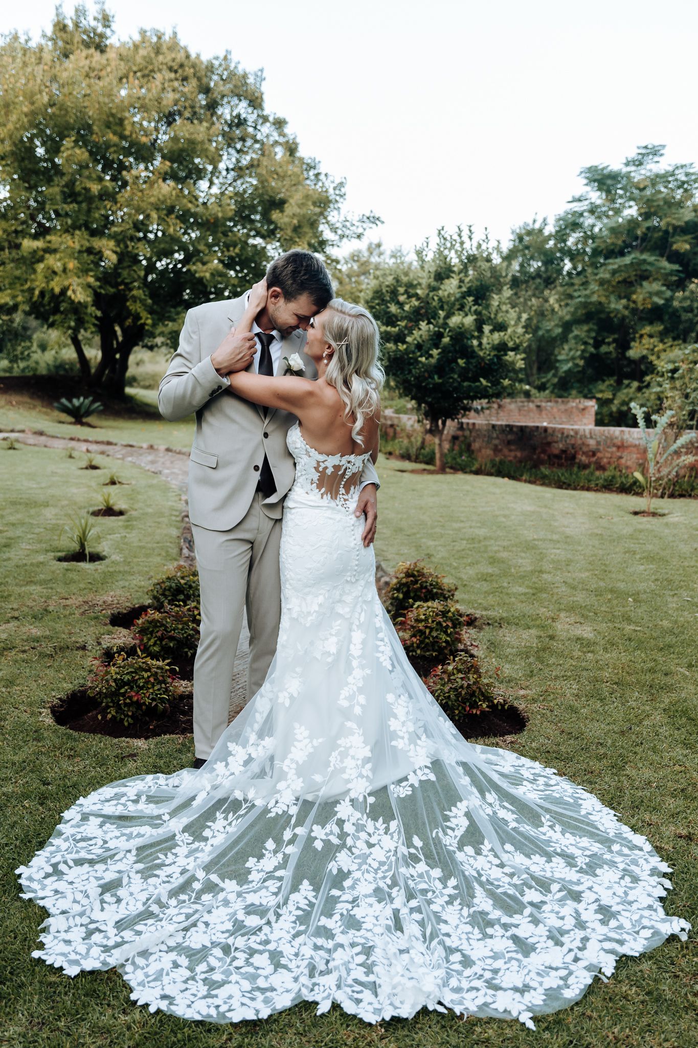 Elegant wedding couple embracing outdoors with bride's detailed lace train spread on lawn.