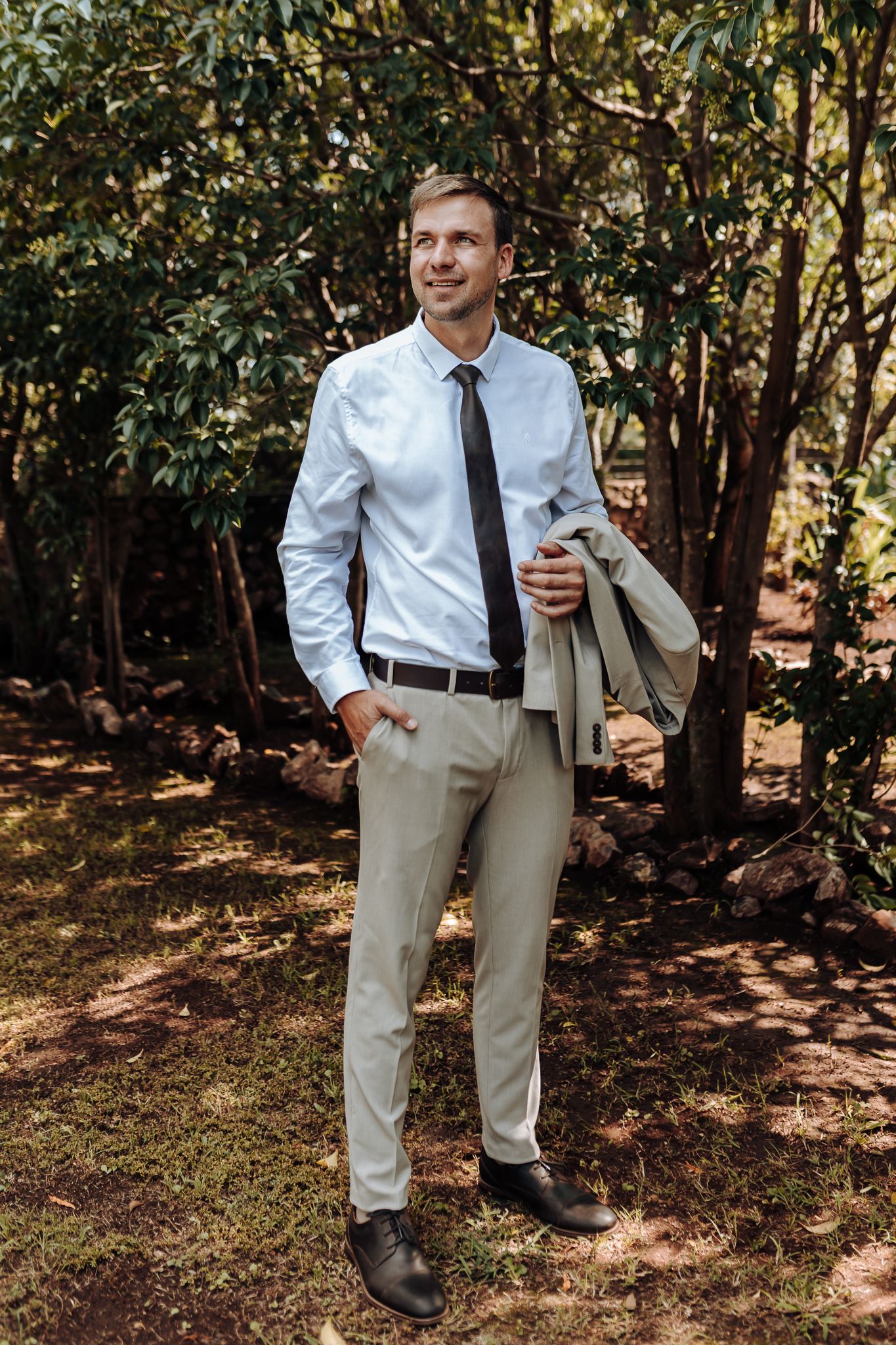 Full-length portrait of man in business attire holding jacket against wooded background.