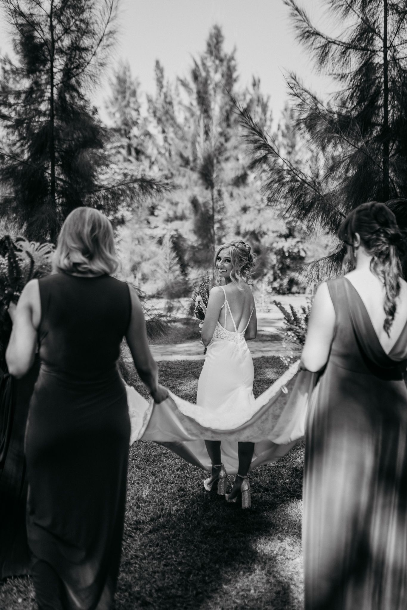 Black and white photo of wedding party helping with dress train in forest setting.