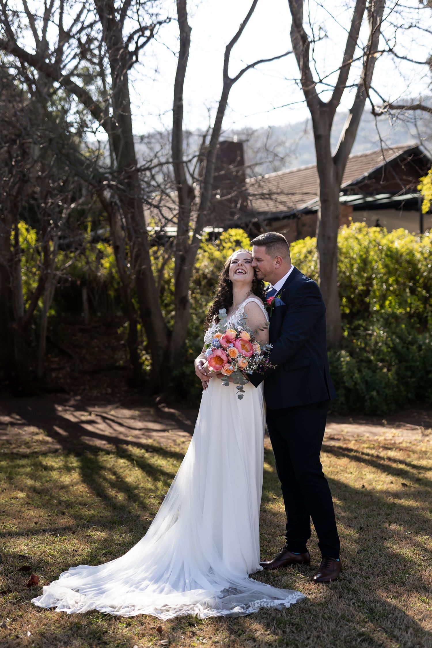 A couple shares a romantic moment in a garden setting with sunlit trees and a flowing white wedding dress.