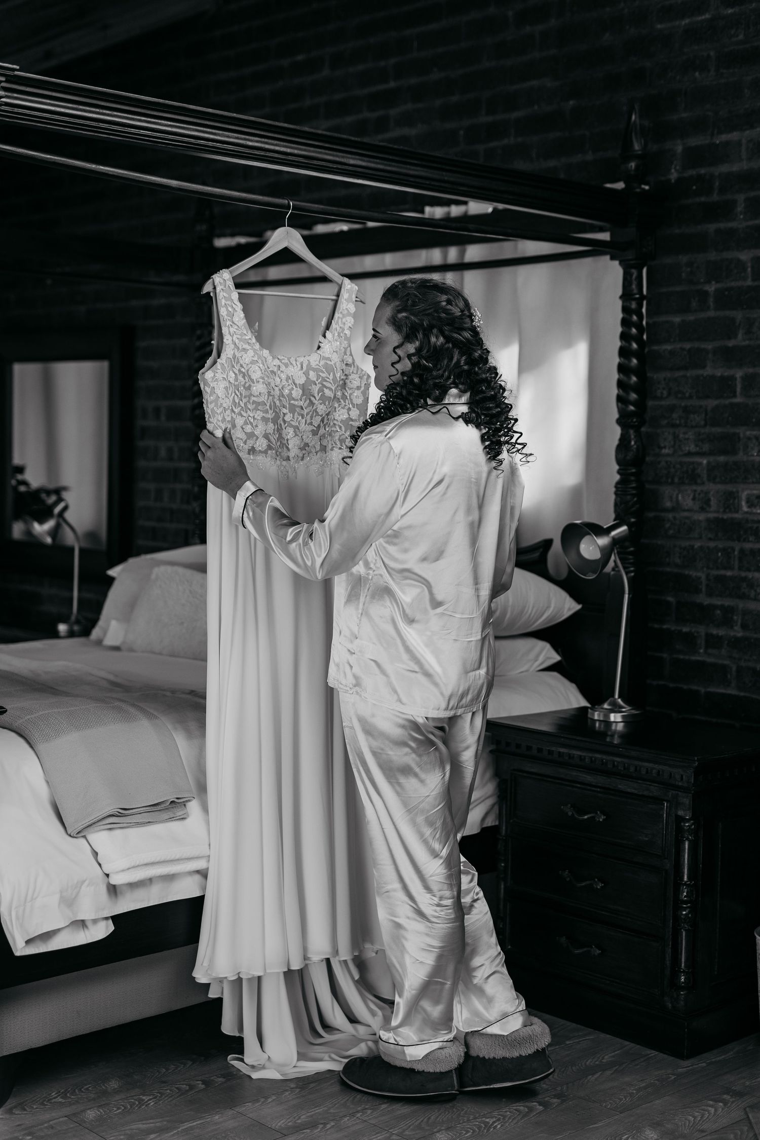 Black and white photo of a flowing wedding dress hanging on display next to someone in white clothing in a bedroom.