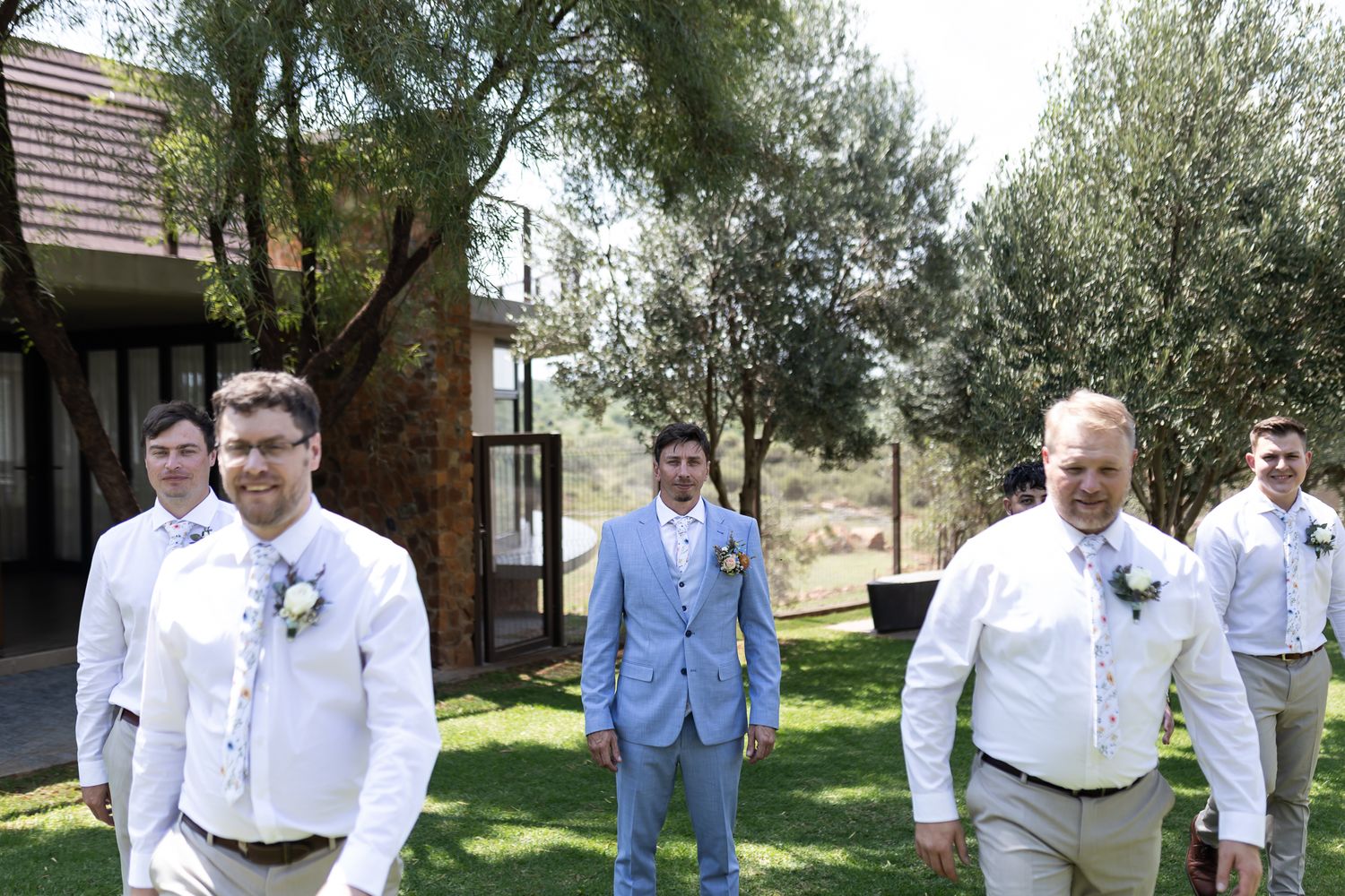 Wedding party in white shirts and light suits pose outdoors with one member wearing powder blue.