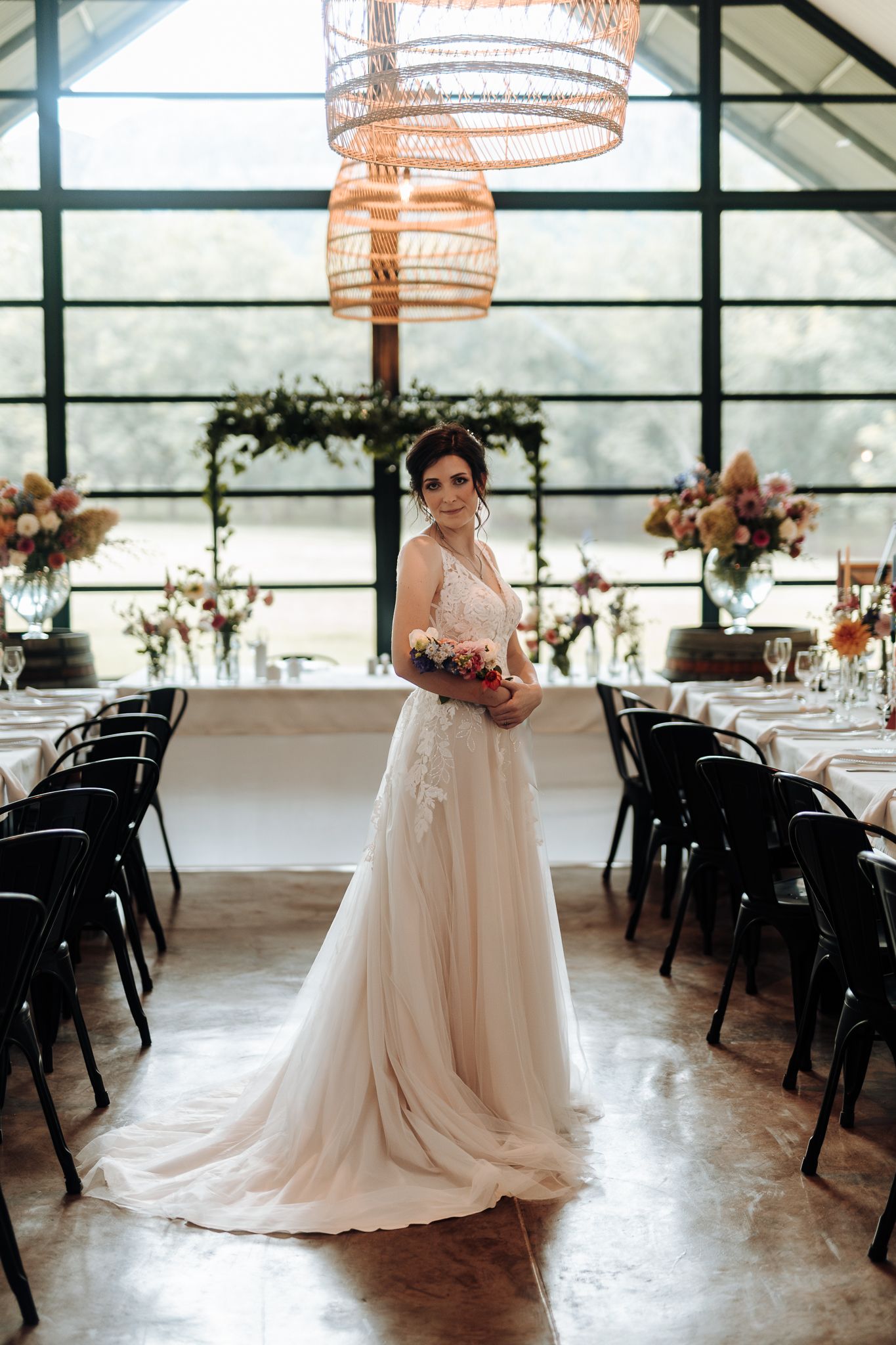 Bride in flowing white gown stands gracefully in sunlit venue with modern chandelier and table settings.
