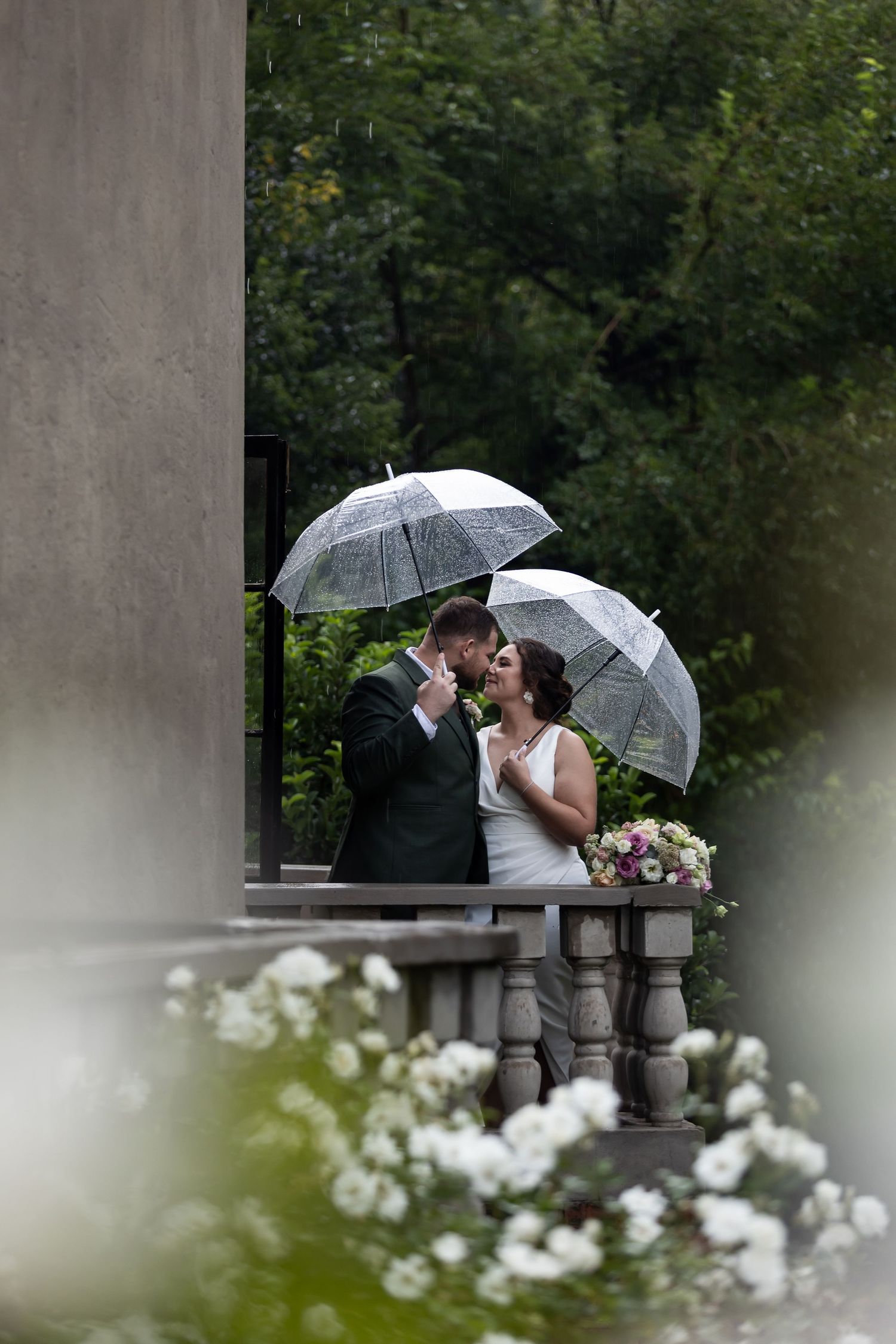 Couple shares a tender moment under clear umbrellas on a rainy wedding day with white flowers in foreground.