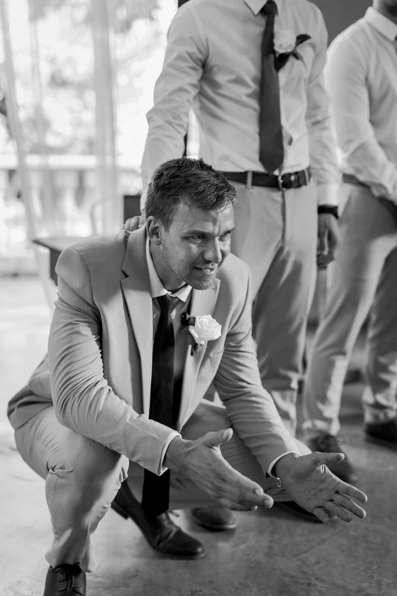 Groom in light gray suit crouches down with arms outstretched during wedding celebration.
