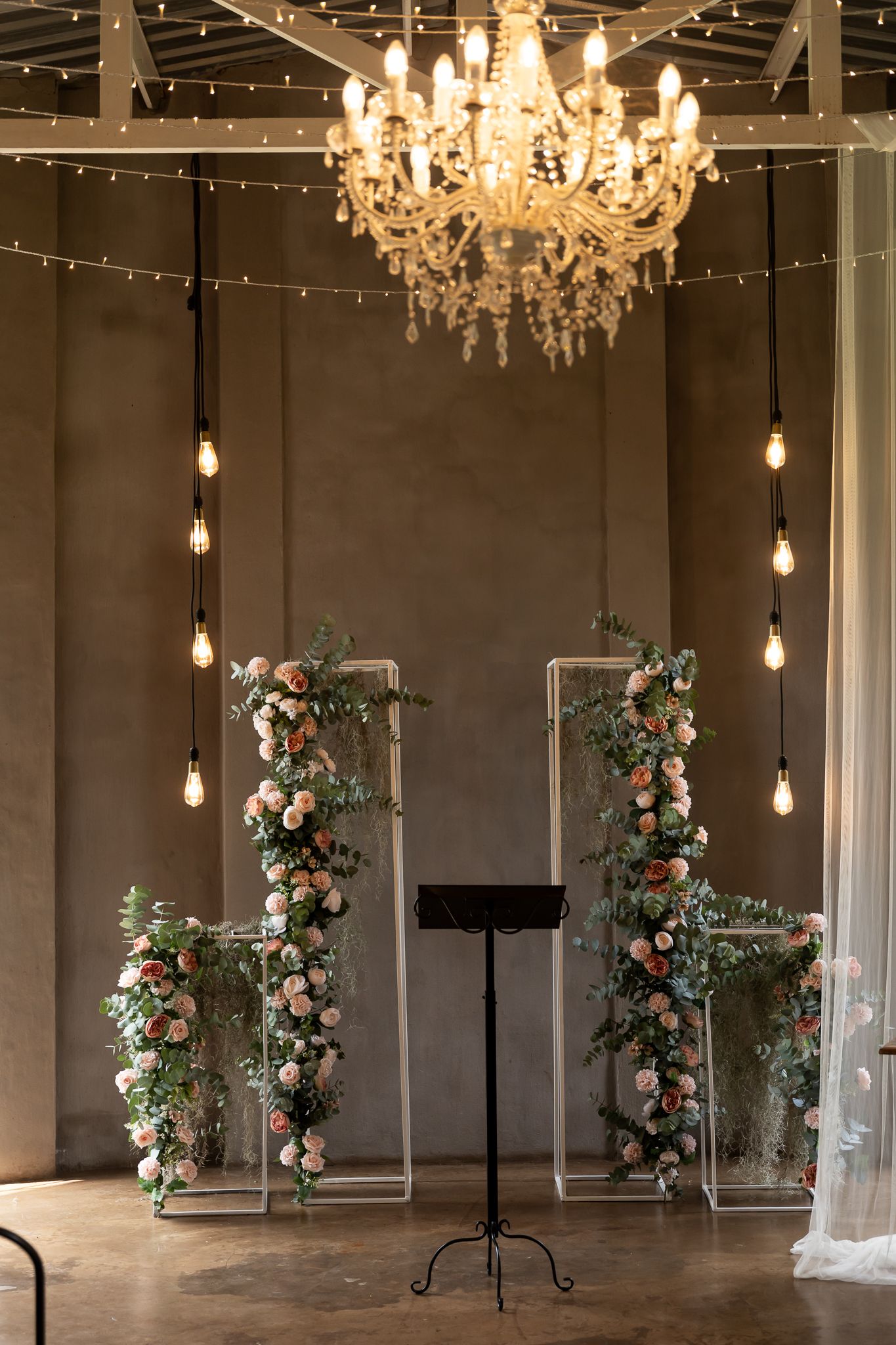 Romantic ceremony backdrop with floral arrangements on pedestals under a crystal chandelier and string lights.