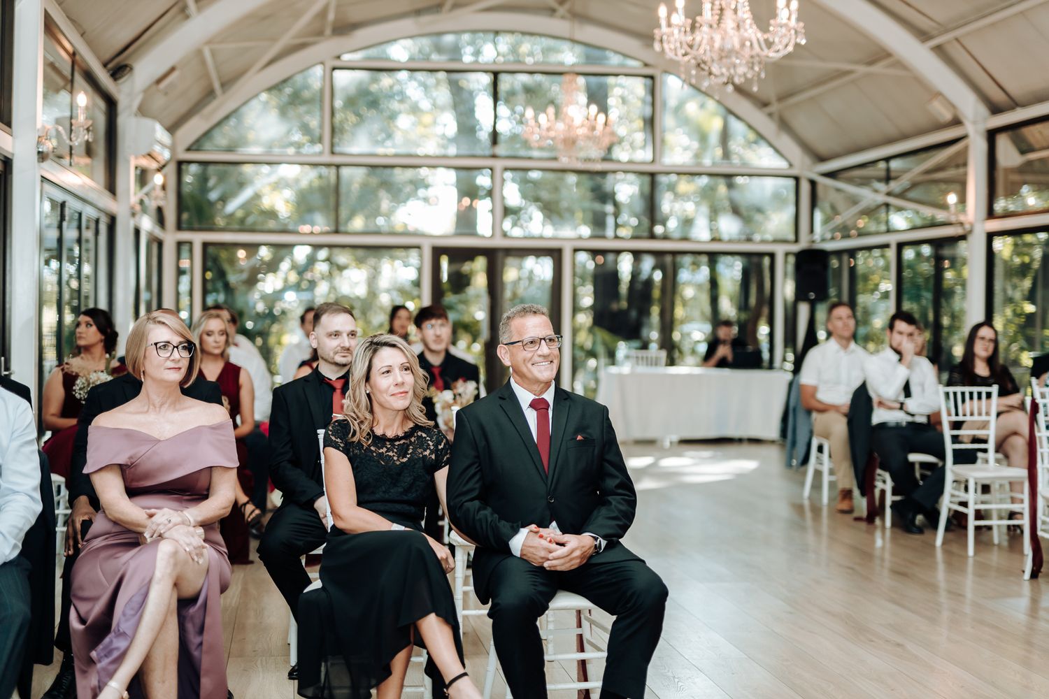 Wedding guests seated in elegant venue with crystal chandeliers and arched windows.