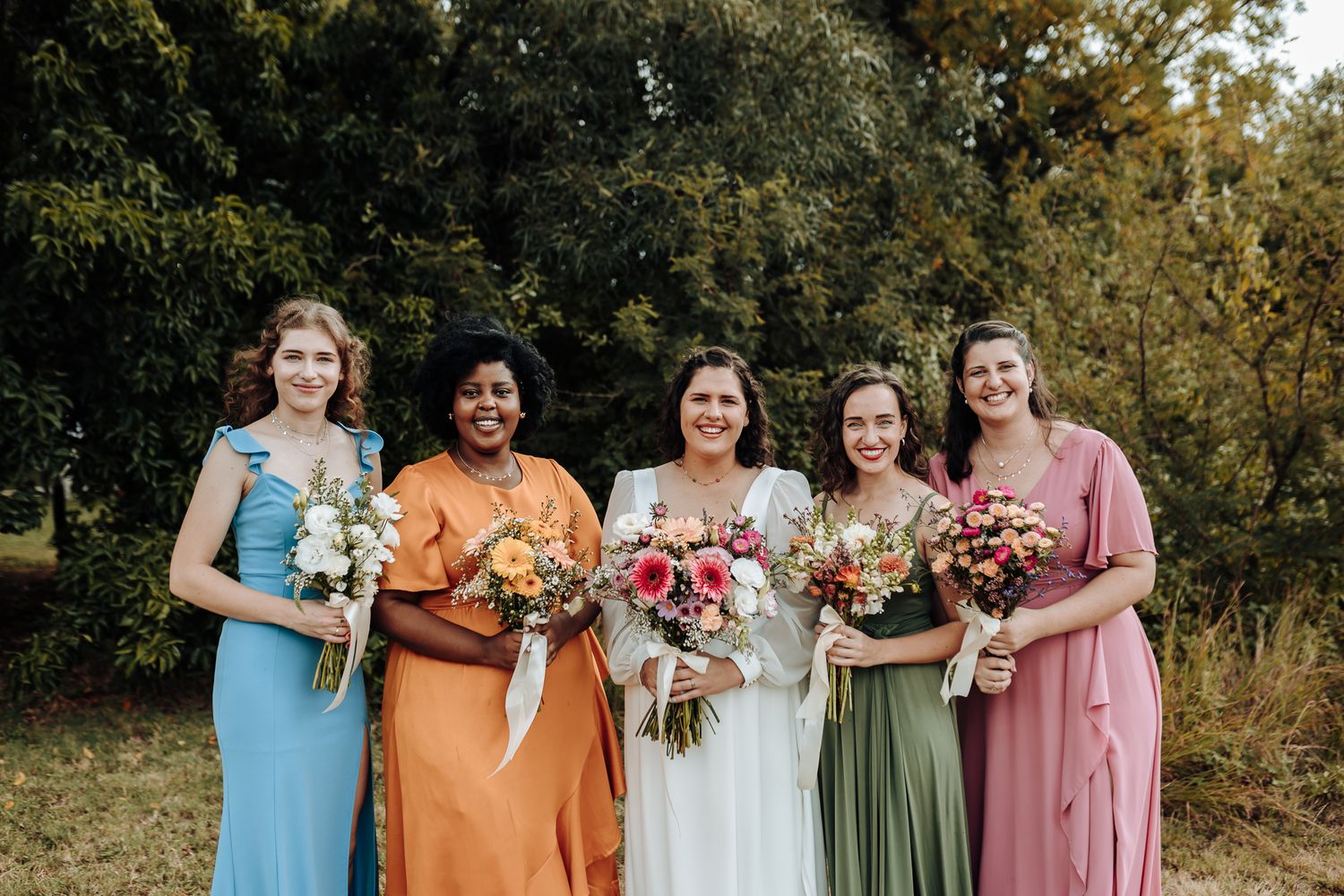 Bridesmaids in colorful dresses of blue, orange, sage green and pink holding bouquets outdoors.