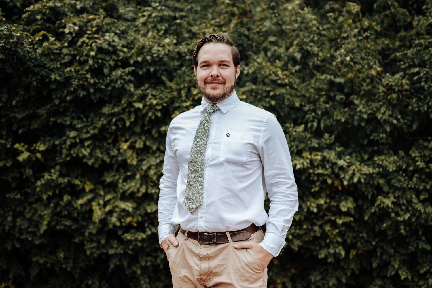 Groomsman in white dress shirt and olive tie smiles against garden foliage.