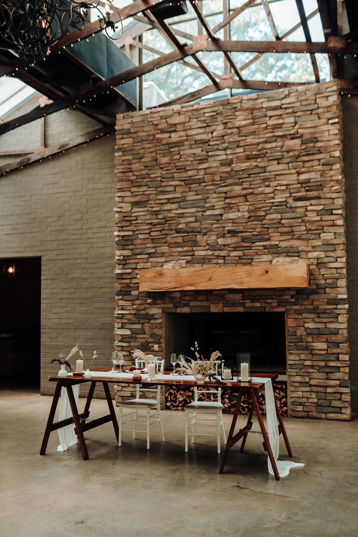 Wedding reception venue with stone fireplace, wooden tables and white chairs under glass ceiling.