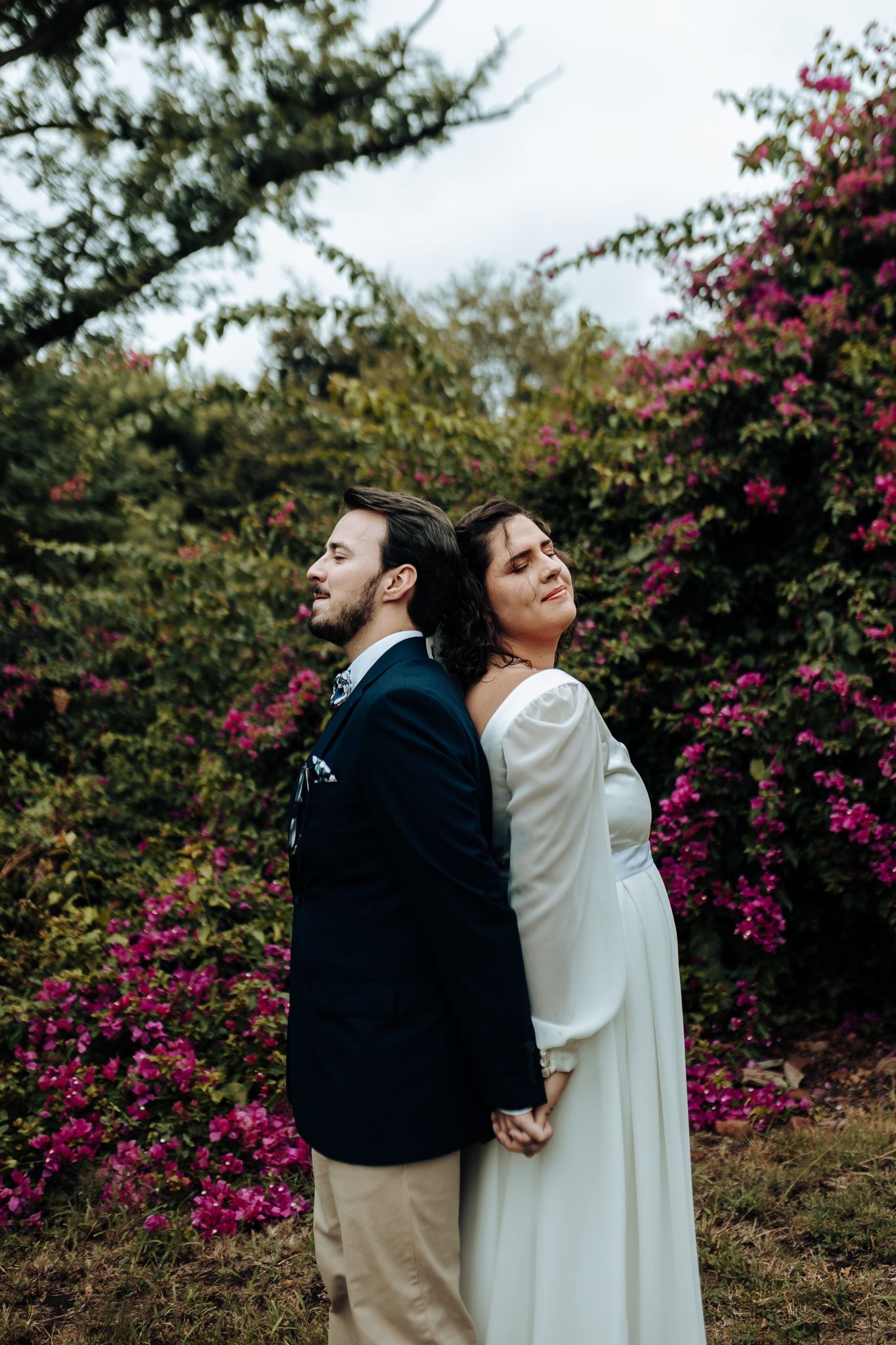 Wedding couple stands back to back holding hands surrounded by pink flowering bushes.