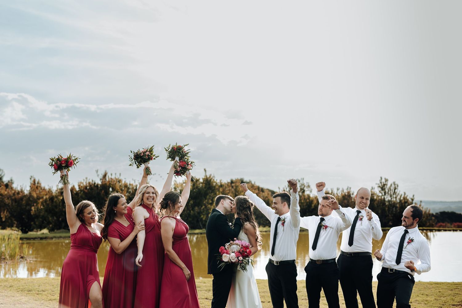 Bridal party celebrates with raised bouquets against natural outdoor setting.