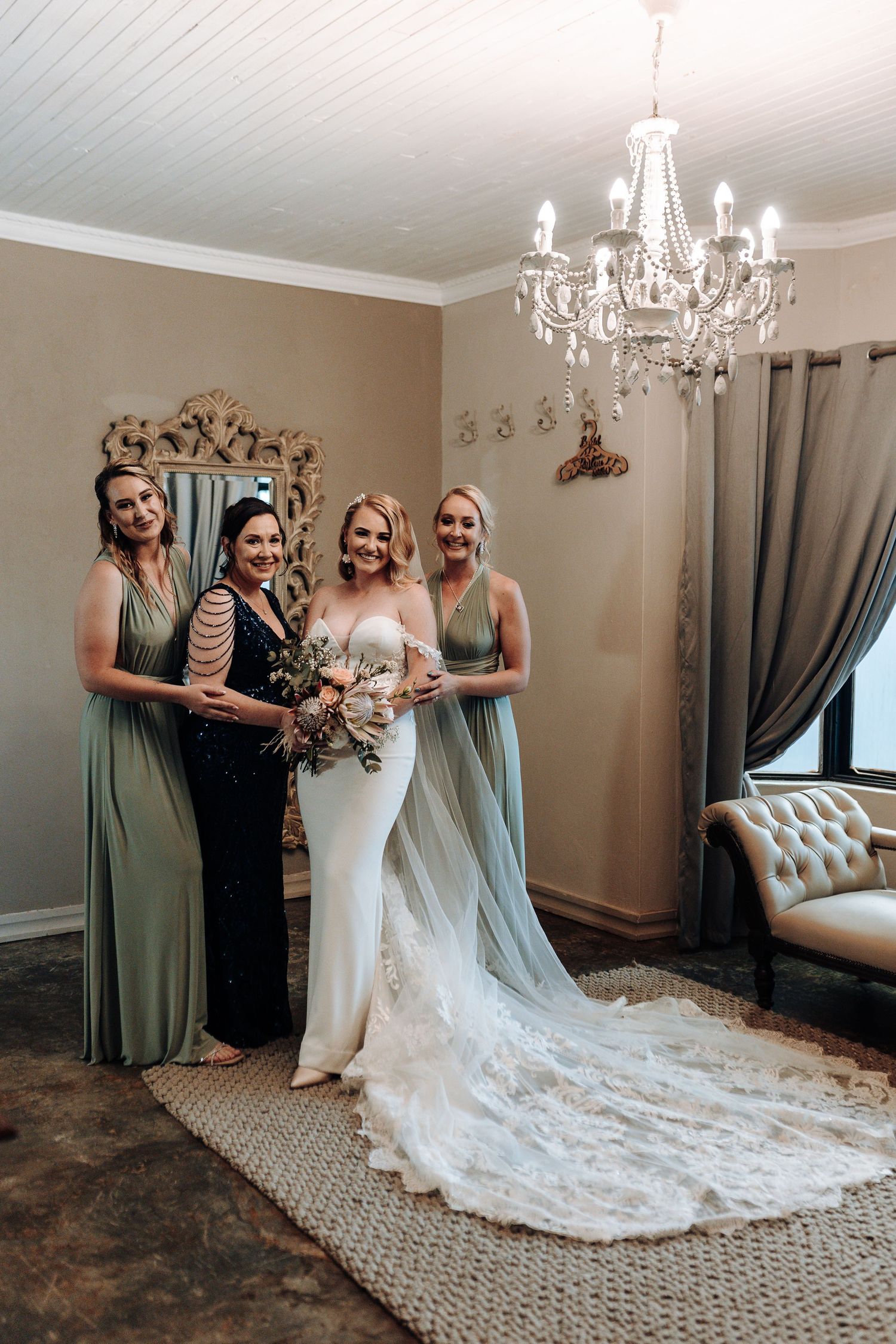 Wedding party poses together under a crystal chandelier with bridesmaids in sage green dresses.