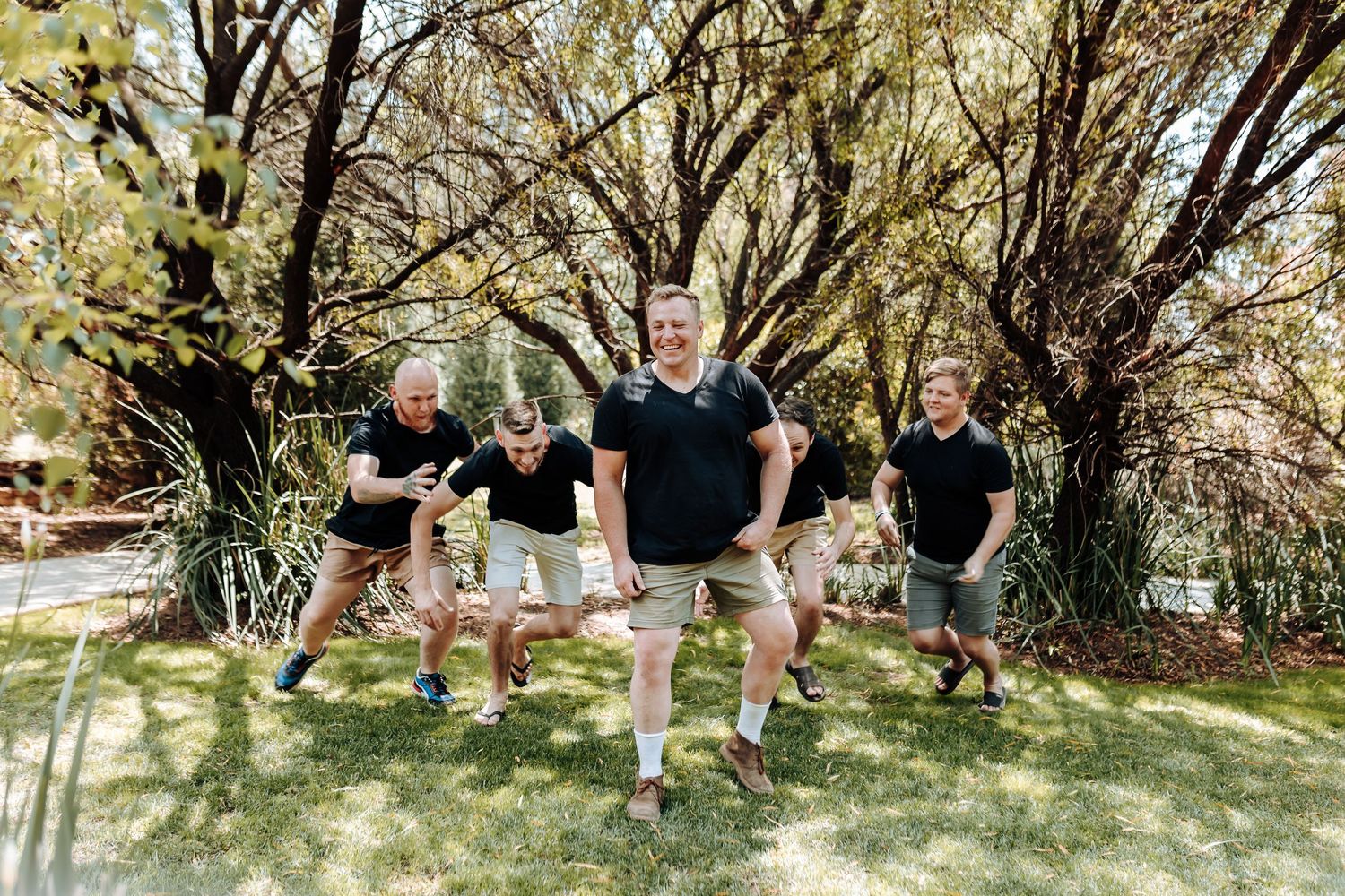 Group poses energetically on grass under trees wearing matching casual attire.