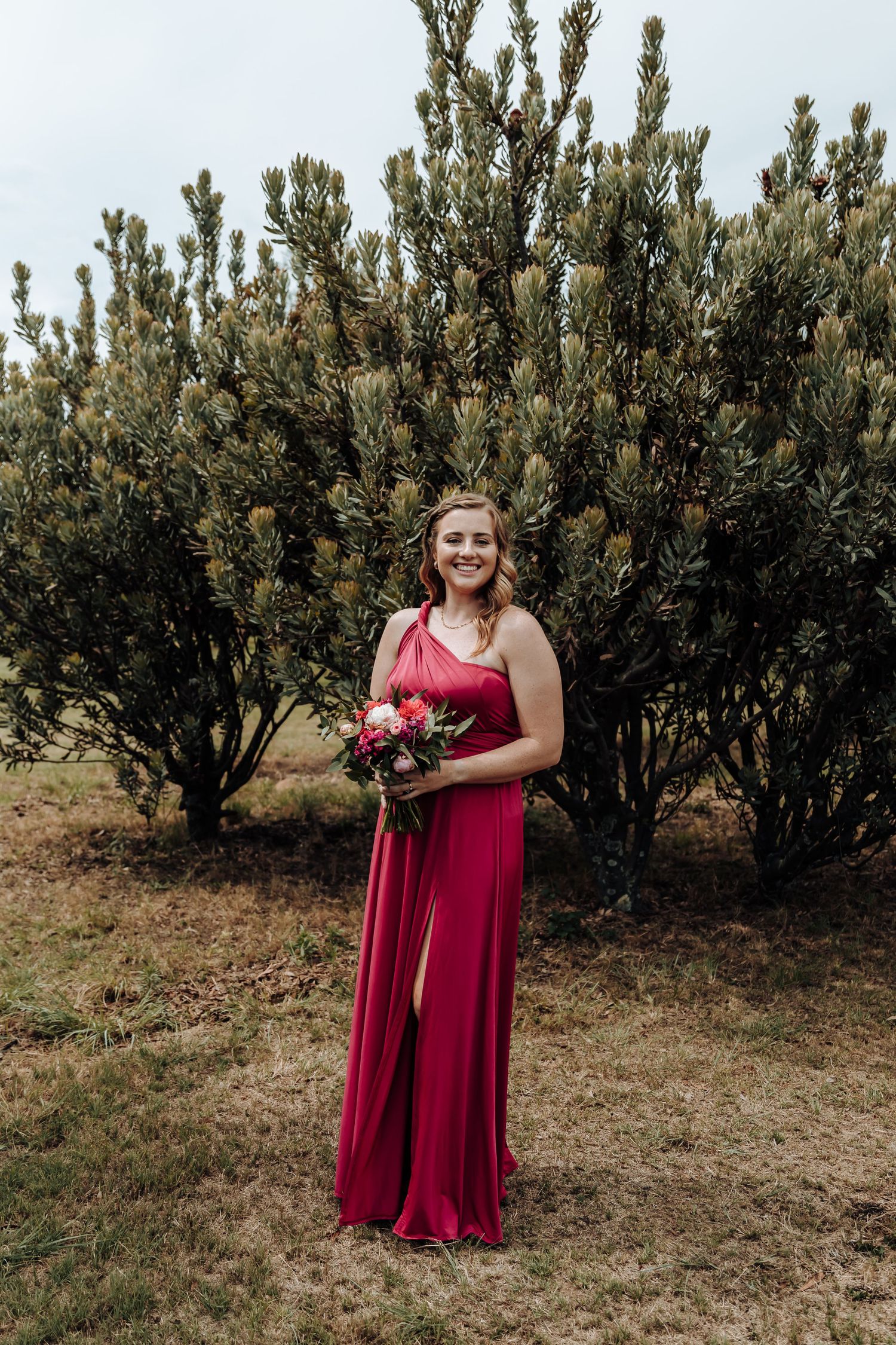 Bridesmaid in flowing red dress poses with bouquet against evergreen shrubs in outdoor setting.