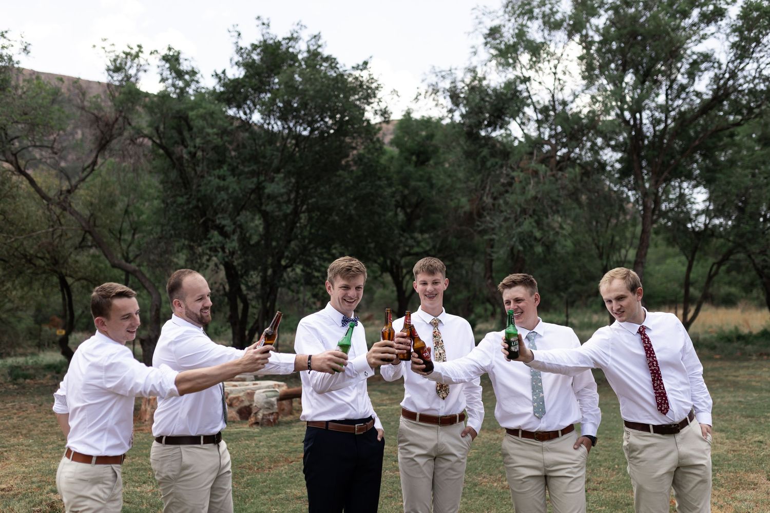 Wedding party raises bottles in a celebratory toast outdoors among trees.