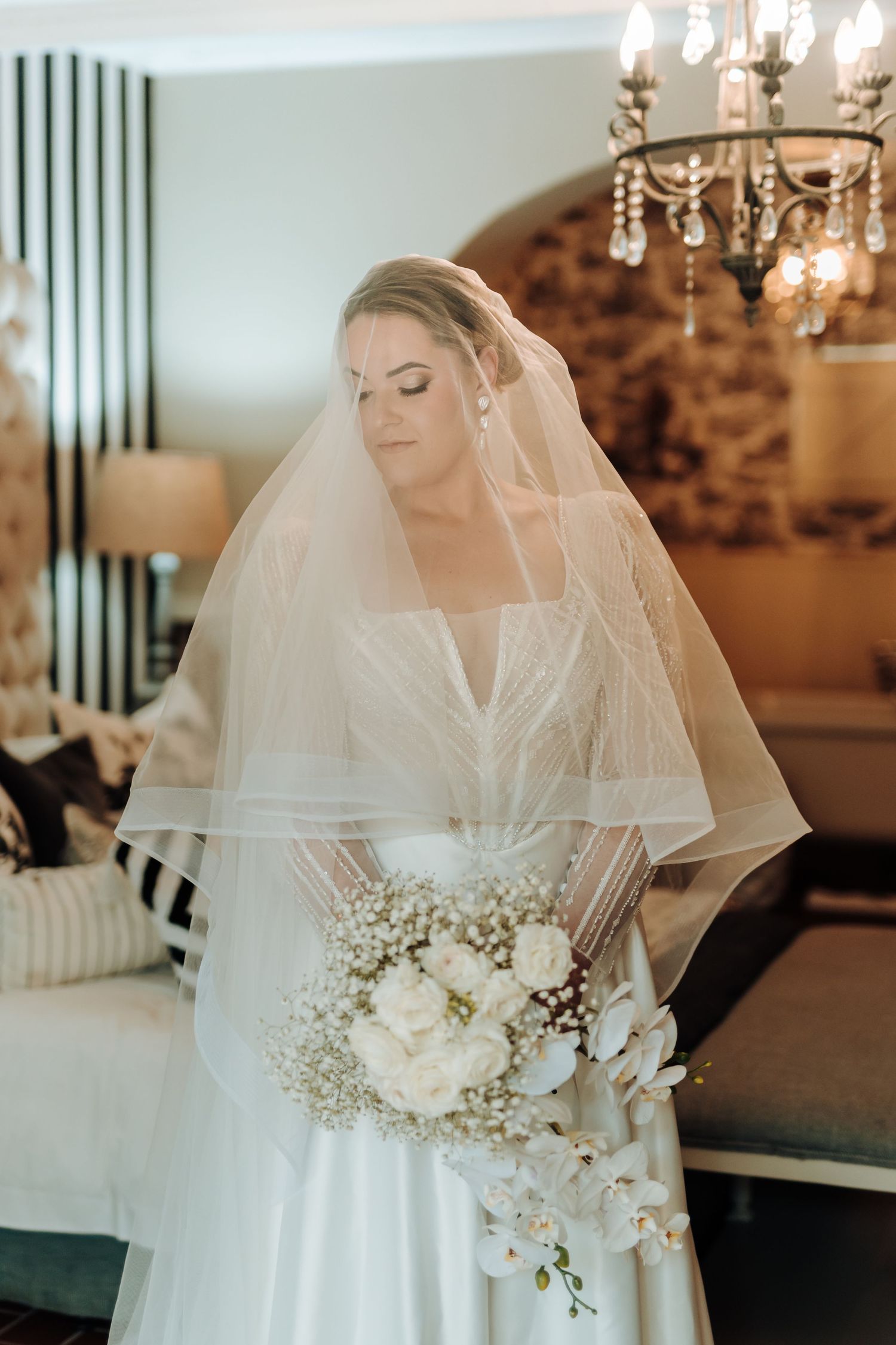 Ethereal bridal portrait with flowing veil and white orchid bouquet under vintage chandelier.