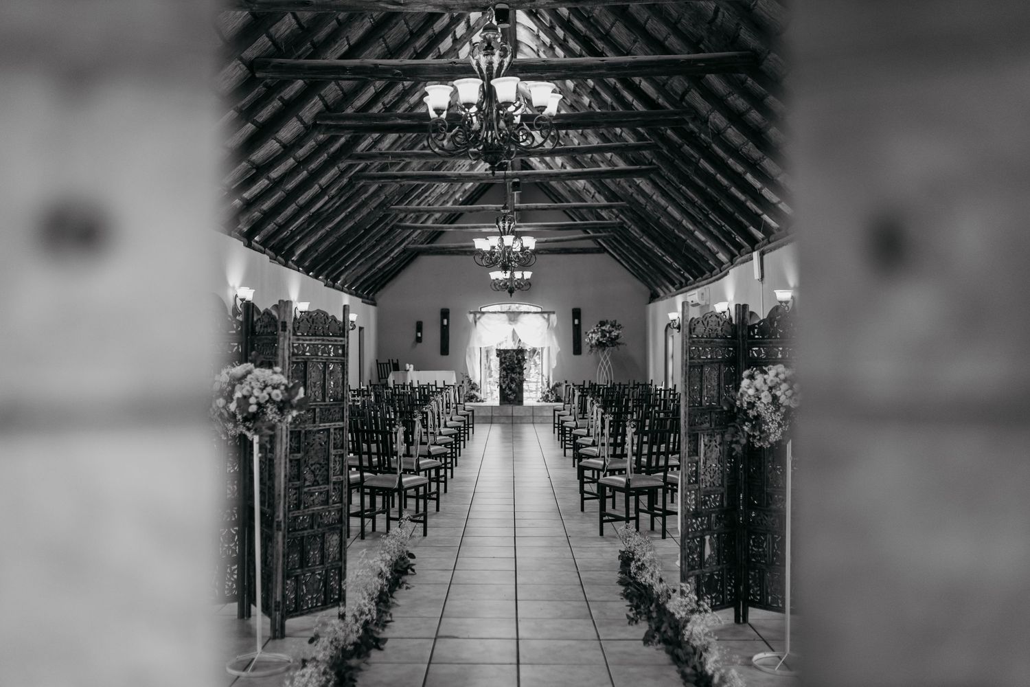 Black and white photo of wedding ceremony aisle with rustic wooden screens and chandeliers.