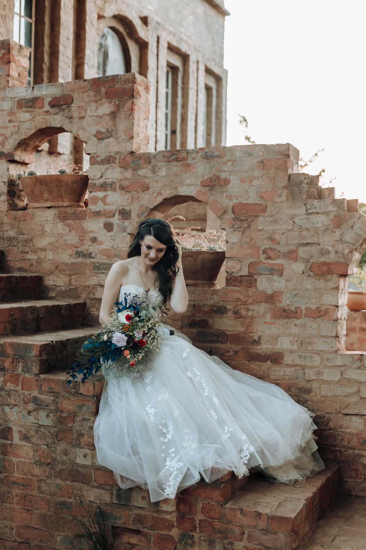 Bridal portrait featuring a lace gown and vibrant bouquet against rustic brick architecture.