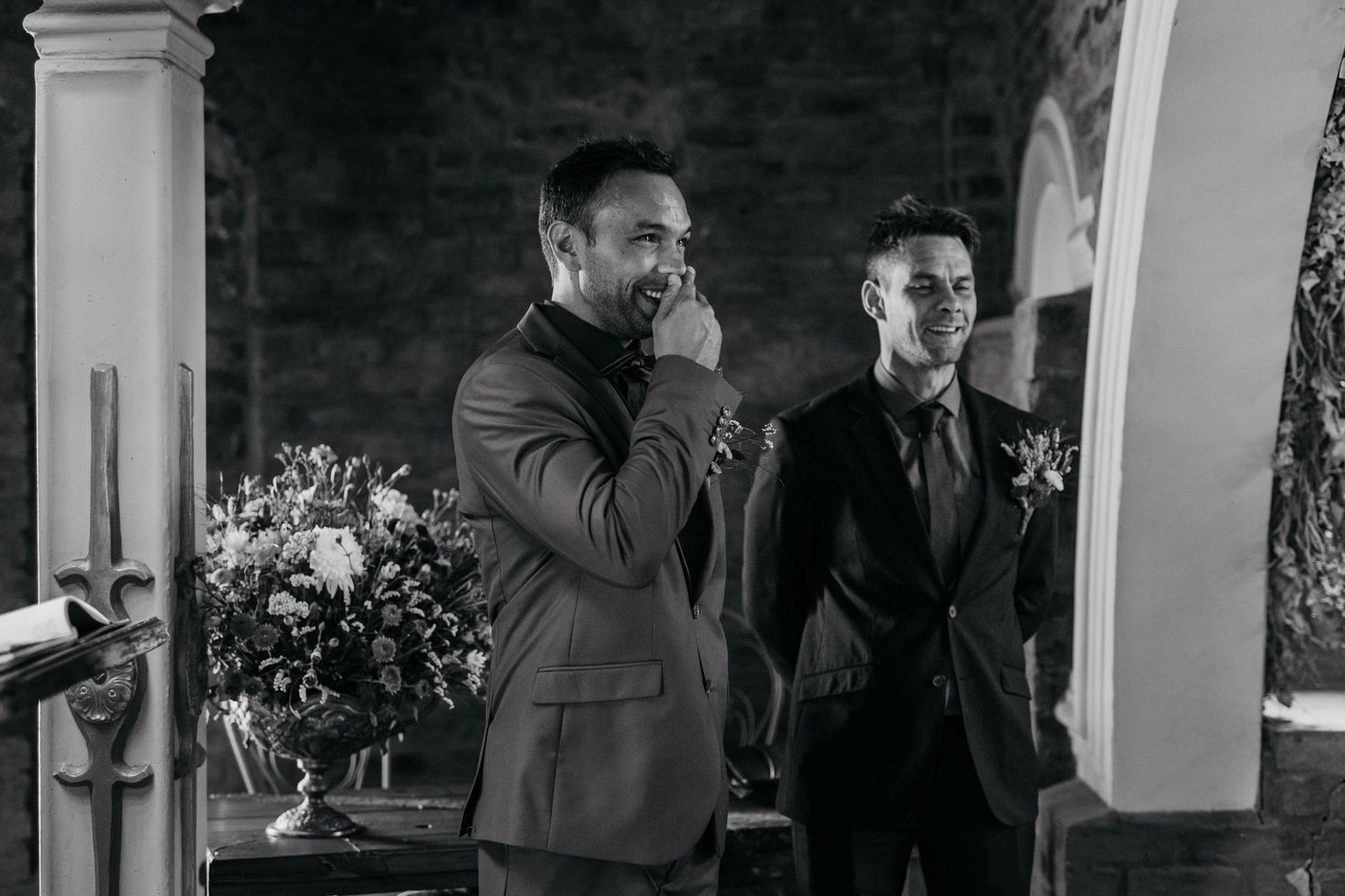 Two wedding guests sharing an emotional moment during ceremony in elegant suits against brick wall backdrop.