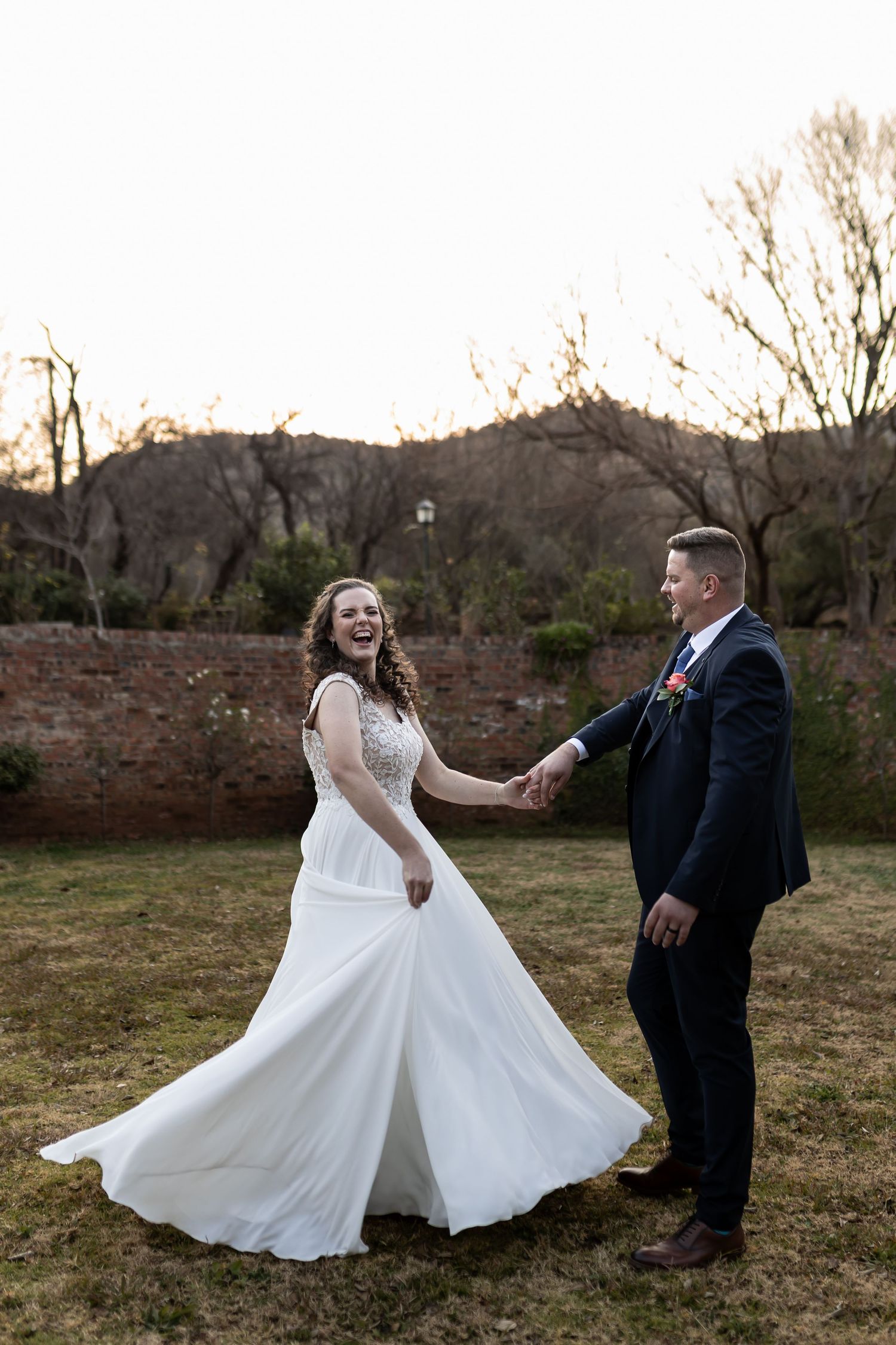 A couple in wedding attire twirls and dances together in a field at sunset creating a romantic portrait series.