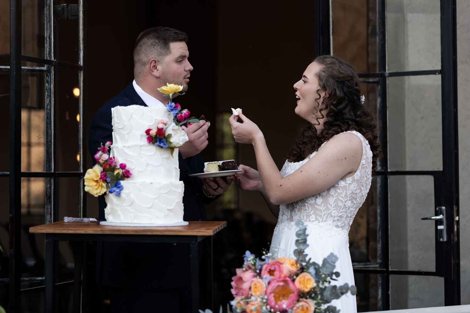 A couple shares their wedding cake during a special moment beside floral decorations at an elegant venue entrance.