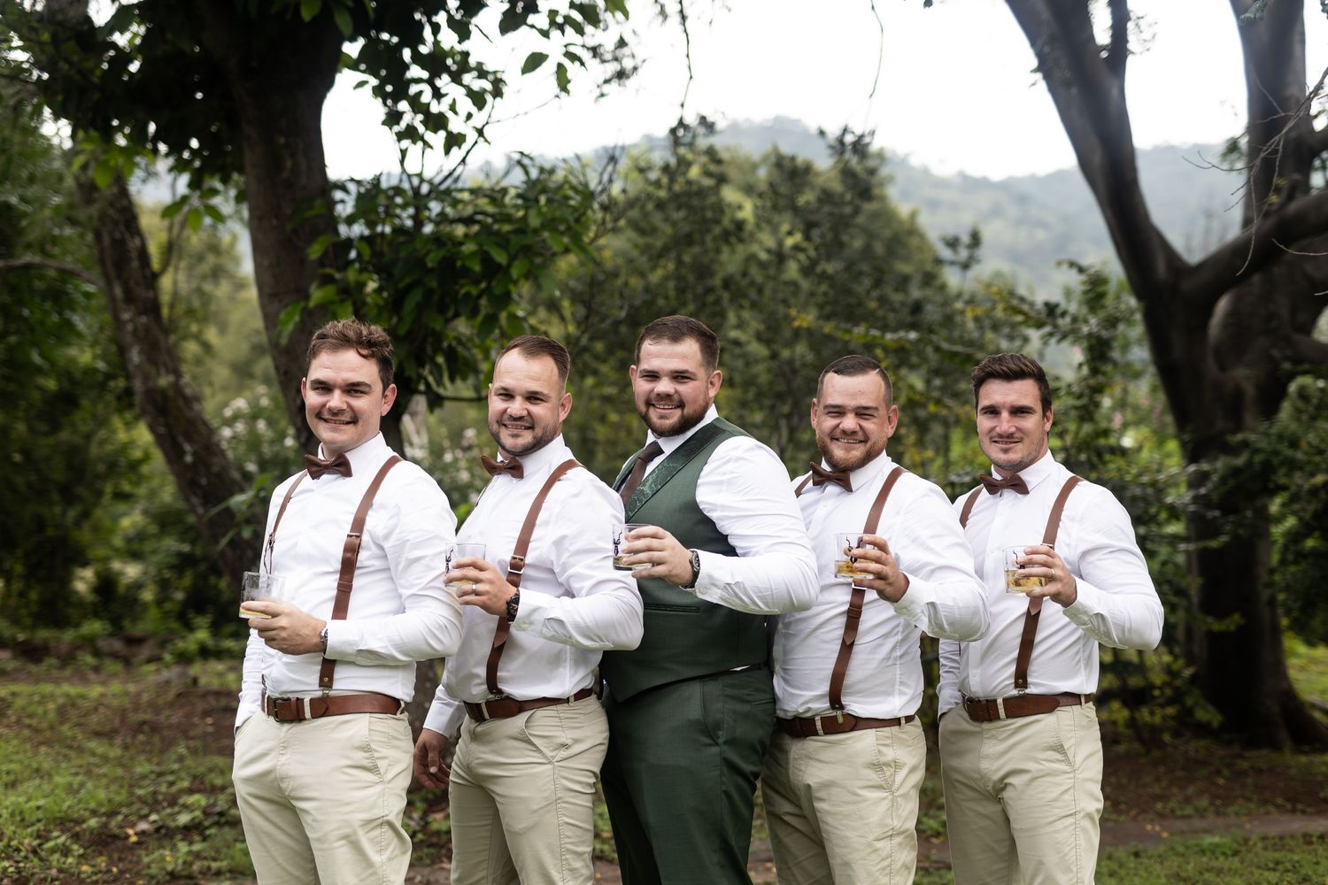 Wedding party in white shirts and suspenders posing together in an outdoor garden setting.