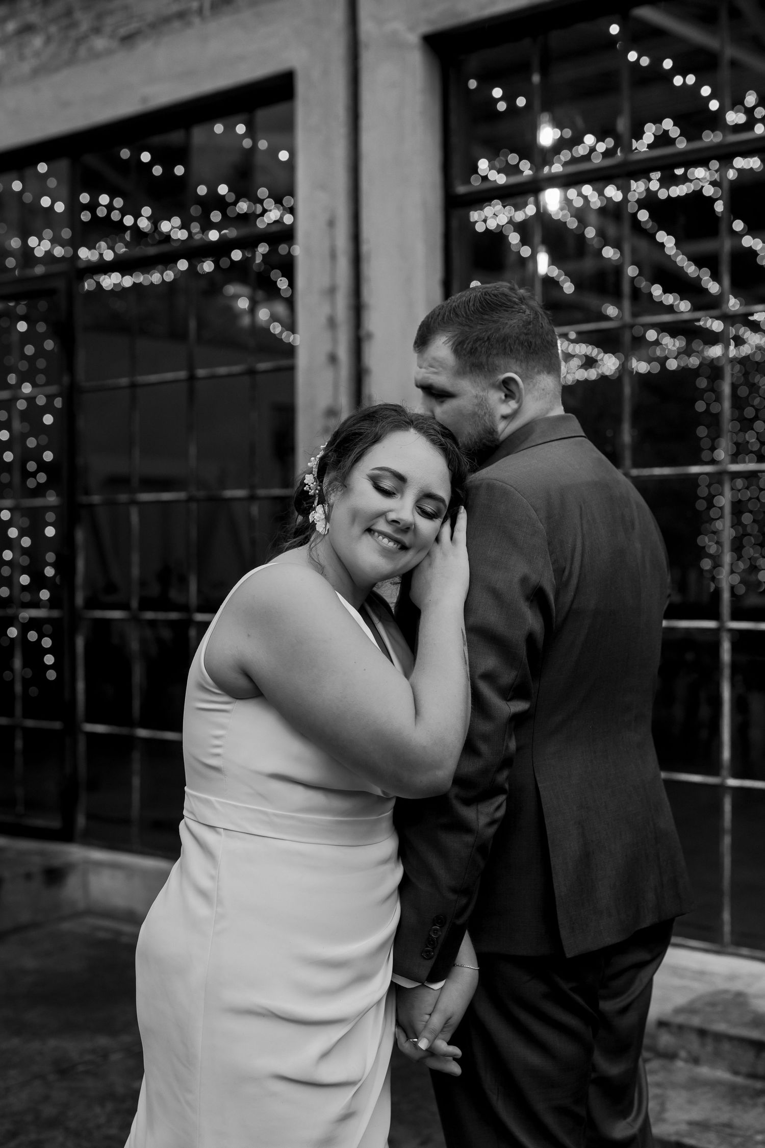 Romantic moment captured in black and white with string lights twinkling in background windows.