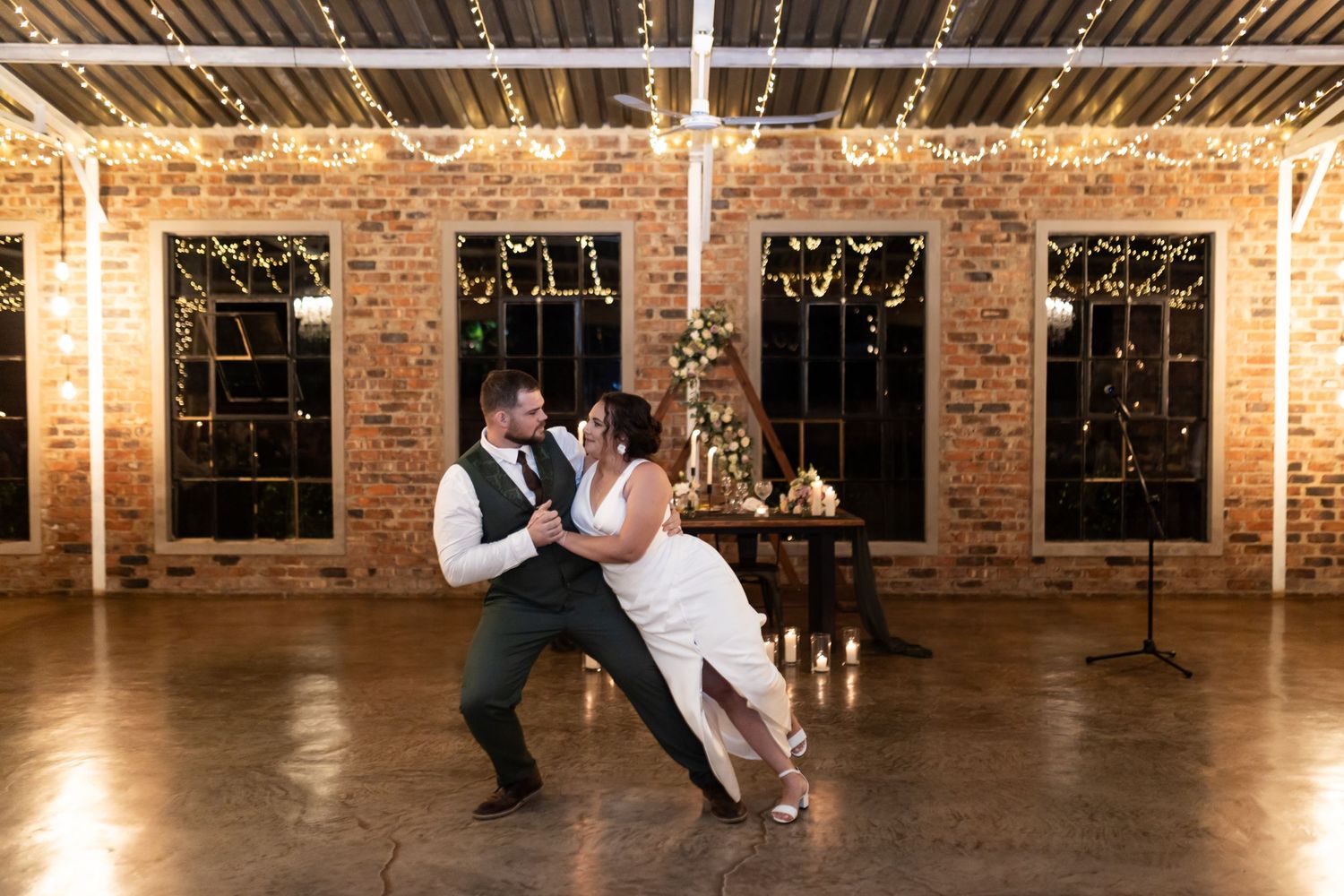 A couple performs a romantic dance dip in an industrial venue with exposed brick walls and string lights.