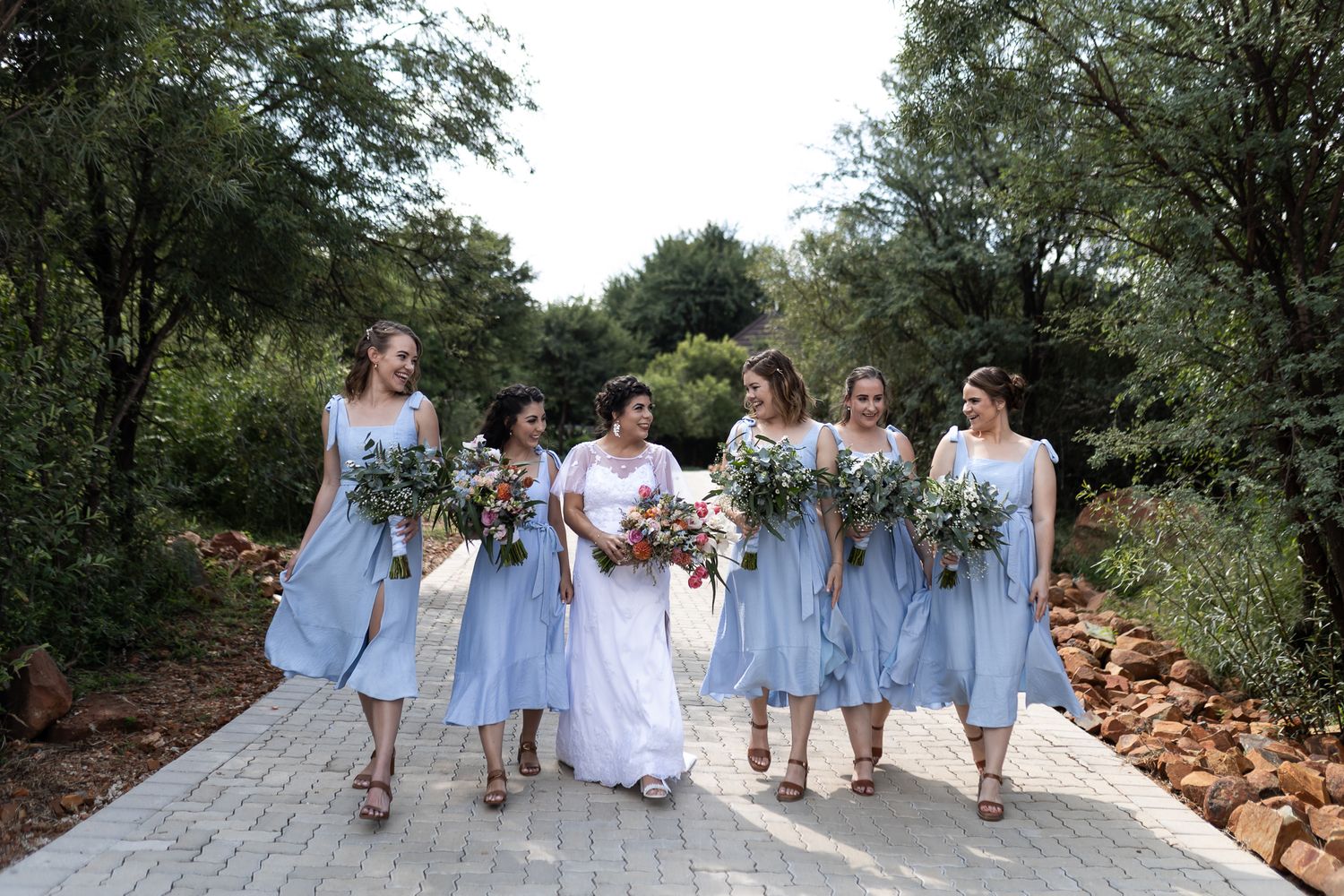 A bridal party in light blue dresses walks together on a stone path surrounded by greenery.