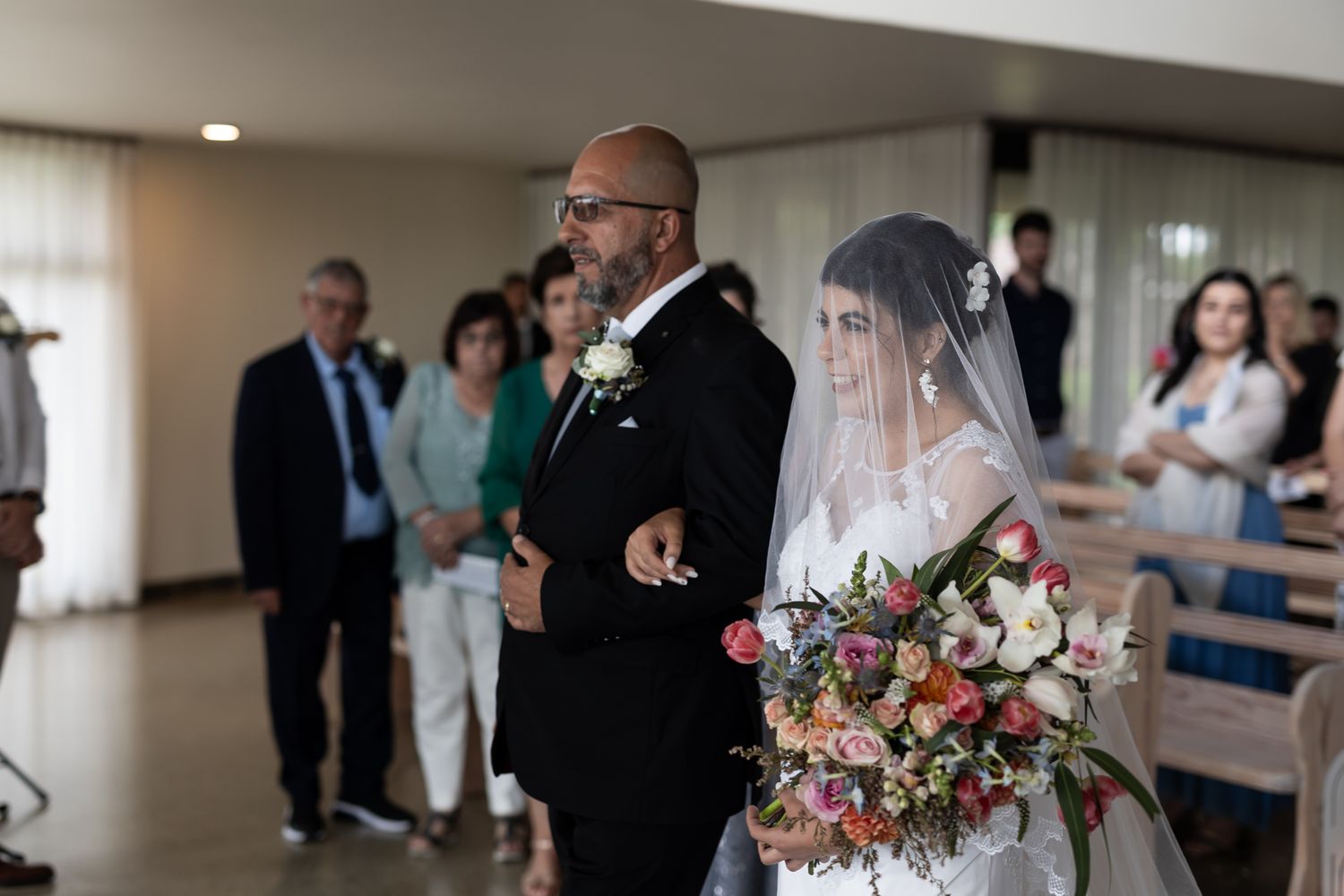 Father walks bride down church aisle carrying vibrant floral bouquet.