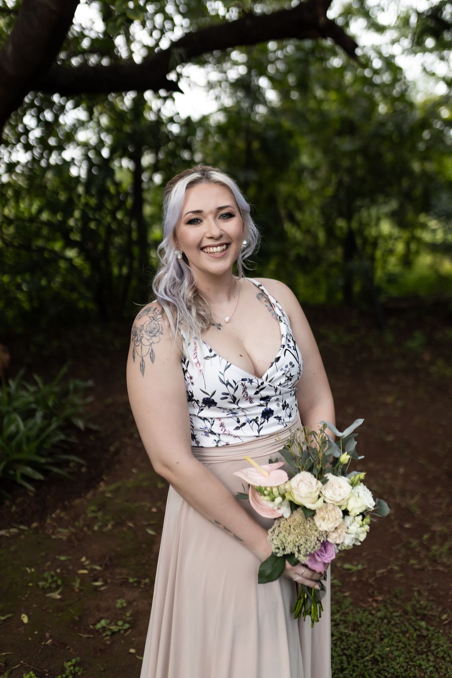 Bridesmaid in floral top and blush skirt holds white and purple bouquet against garden backdrop.